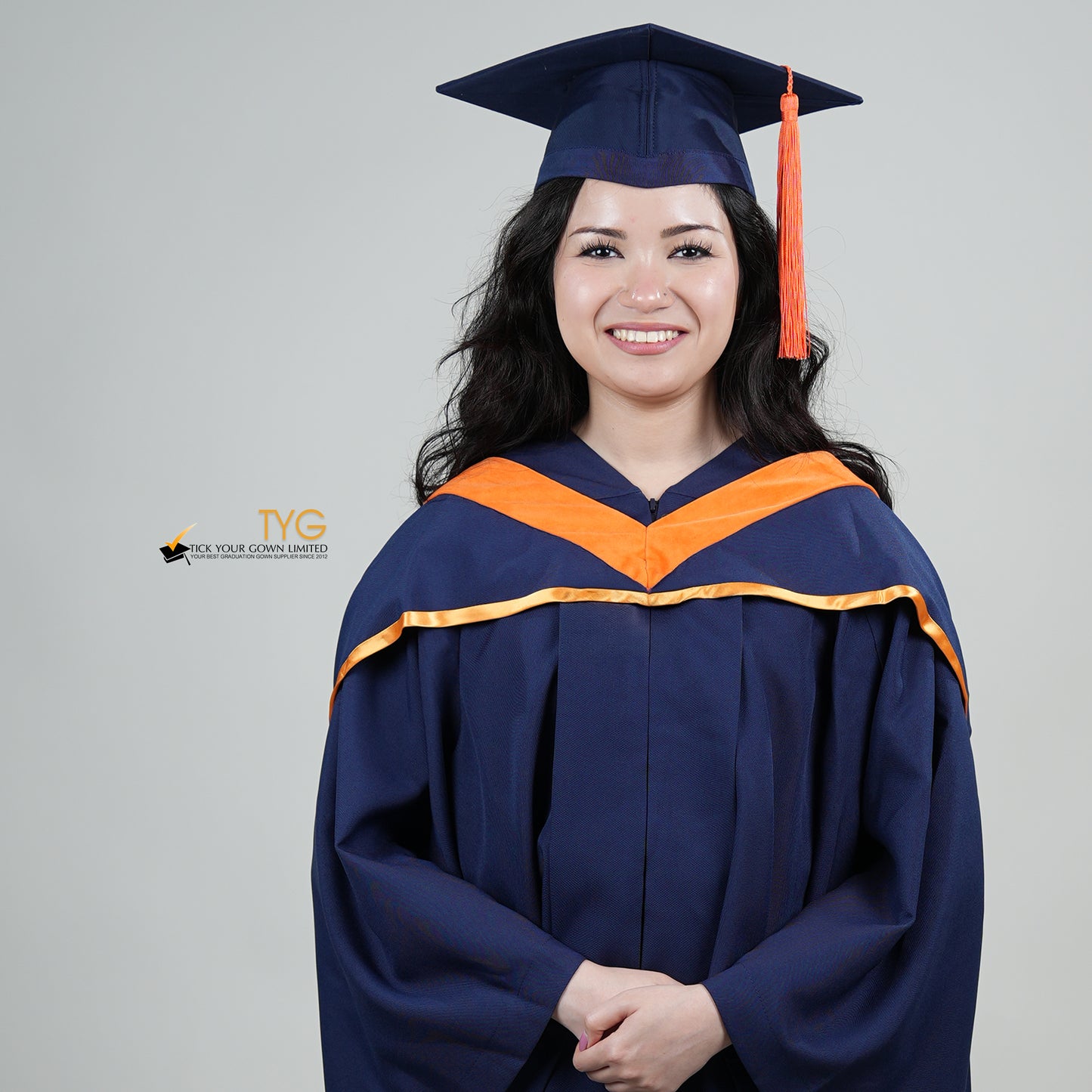 A person in a navy blue graduation gown and an orange hood and with a mortarboard cap with an orange tassel against a gray background.