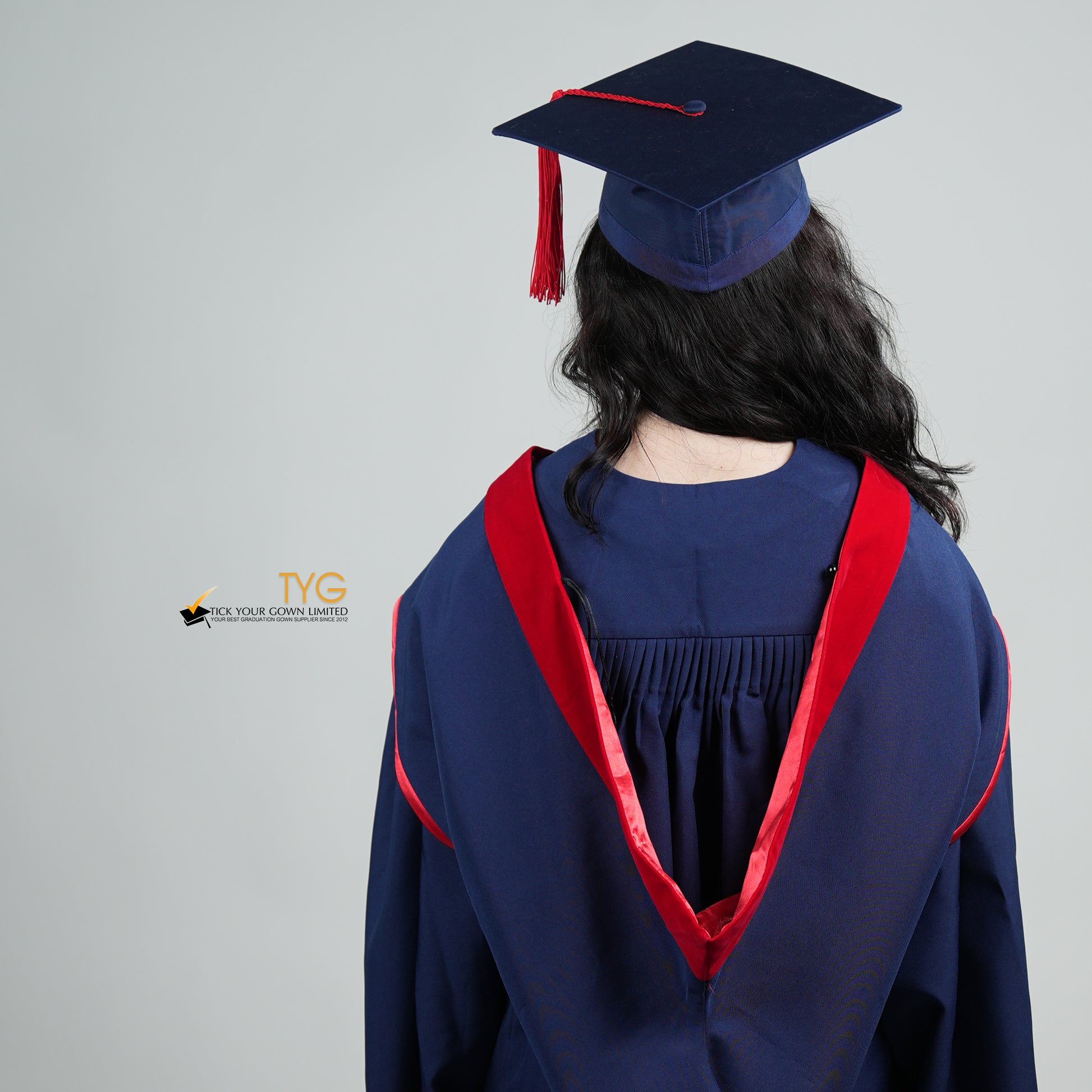 A back view of a person in a navy blue graduation gown and a red hood and with a mortarboard cap with a red tassel against a gray background.