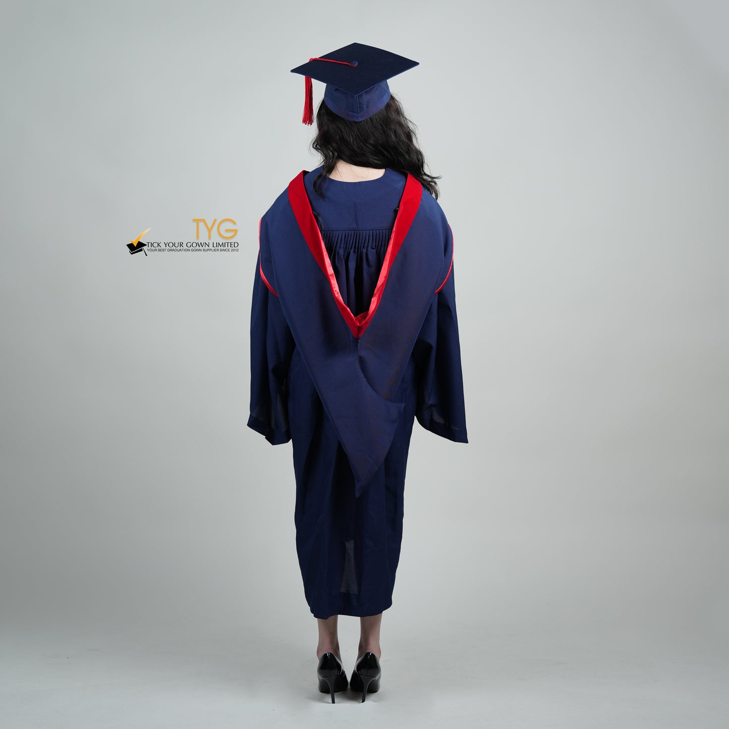 A full body shot of a person in a navy blue graduation gown and a red hood and with a mortarboard cap with a red tassel against a gray background.