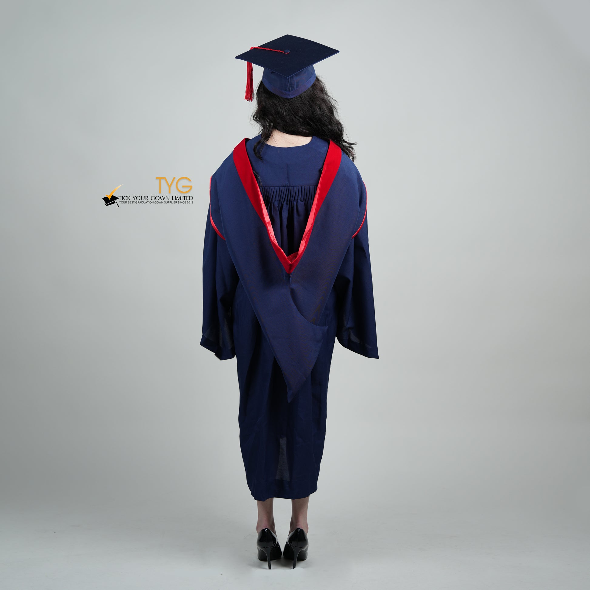 A full body shot of a person in a navy blue graduation gown and a red hood and with a mortarboard cap with a red tassel against a gray background.