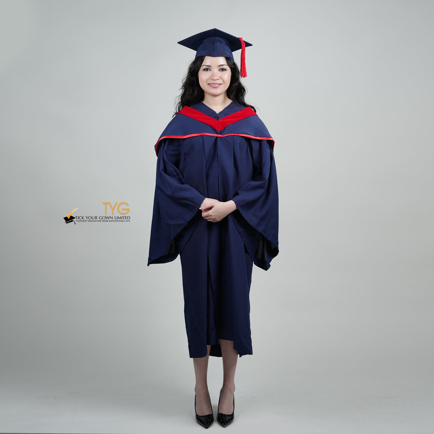 A full body shot of a person in a navy blue graduation gown and a red hood and with a mortarboard cap with a red tassel against a gray background.