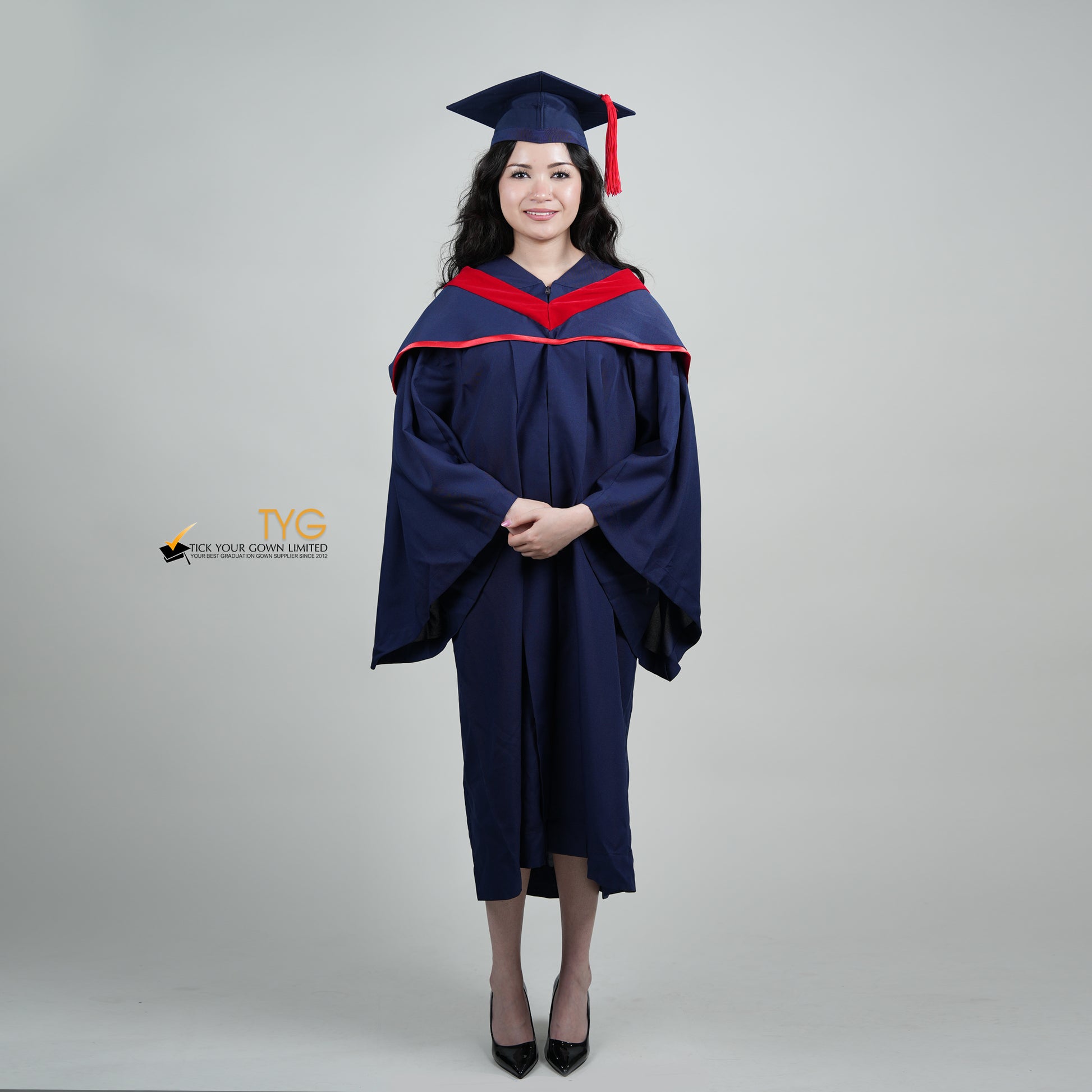 A full body shot of a person in a navy blue graduation gown and a red hood and with a mortarboard cap with a red tassel against a gray background.