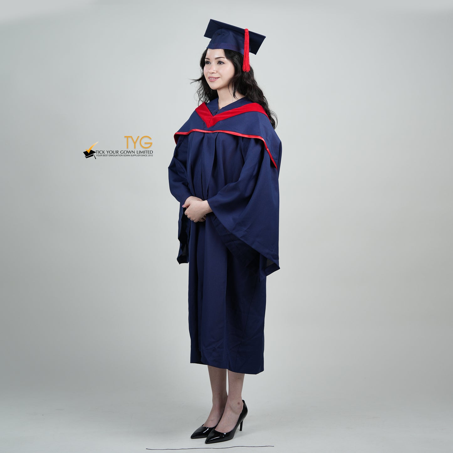 A full body profile view of a person in a navy blue graduation gown and a red hood and with a mortarboard cap with a red tassel against a gray background.