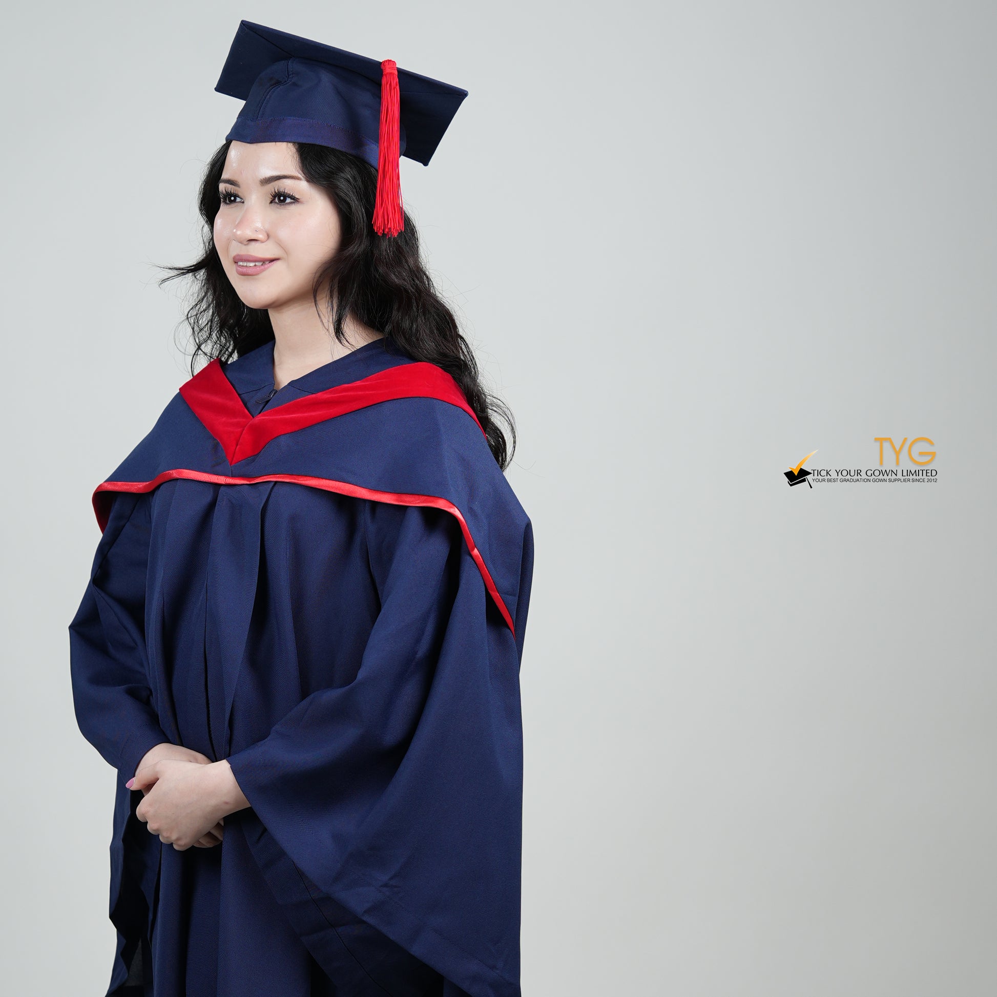 A profile view of a person in a navy blue graduation gown and a red hood and with a mortarboard cap with a red tassel against a gray background.