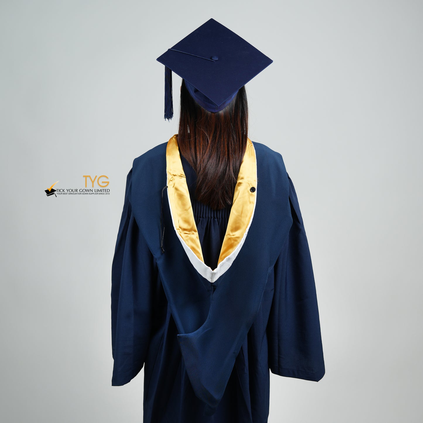A back view of a person in a navy blue graduation gown and a white hood with gold lining and with a mortarboard cap with a white tassel against a gray background.