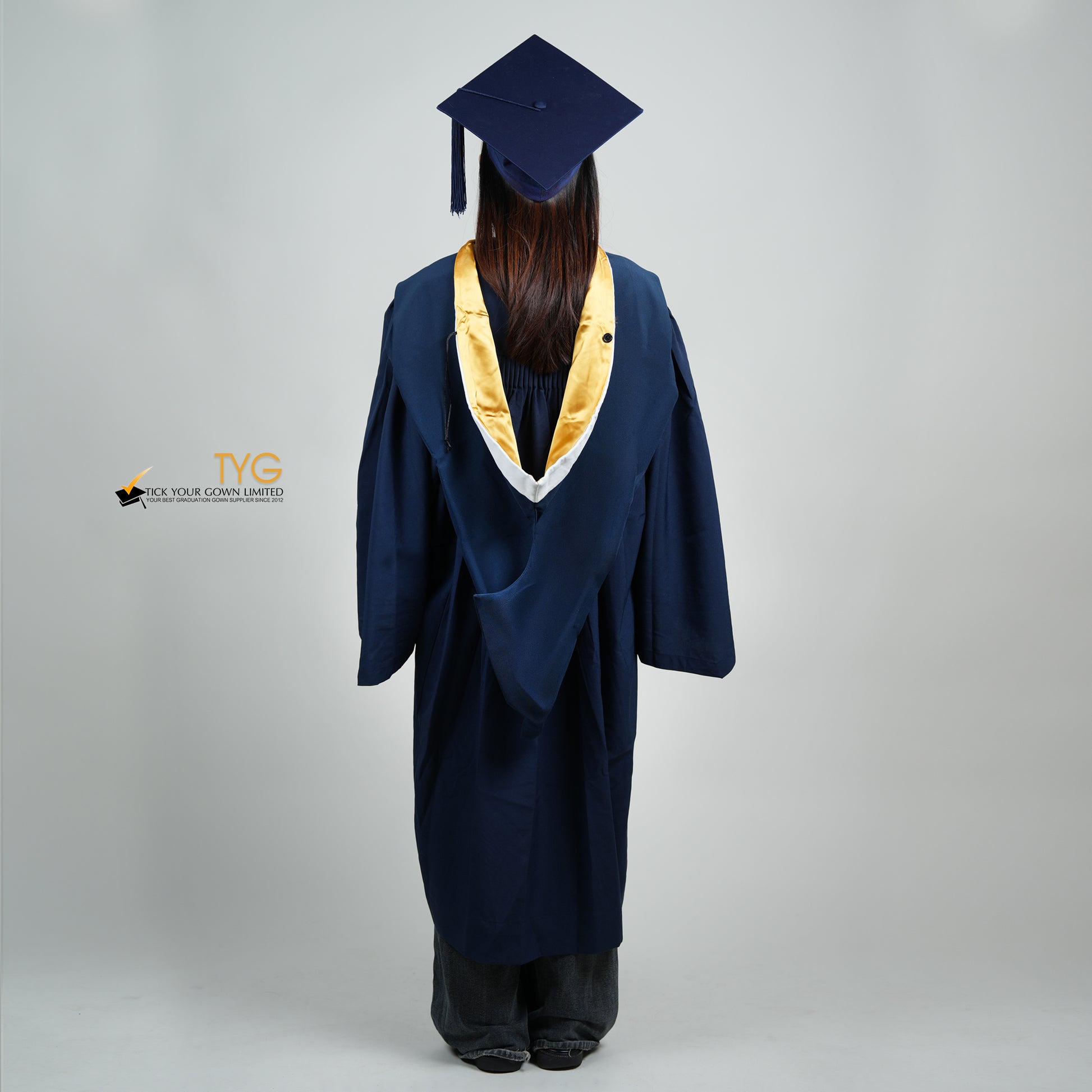 A full body back view of a person in a navy blue graduation gown and a white hood with gold lining and with a mortarboard cap with a white tassel against a gray background.