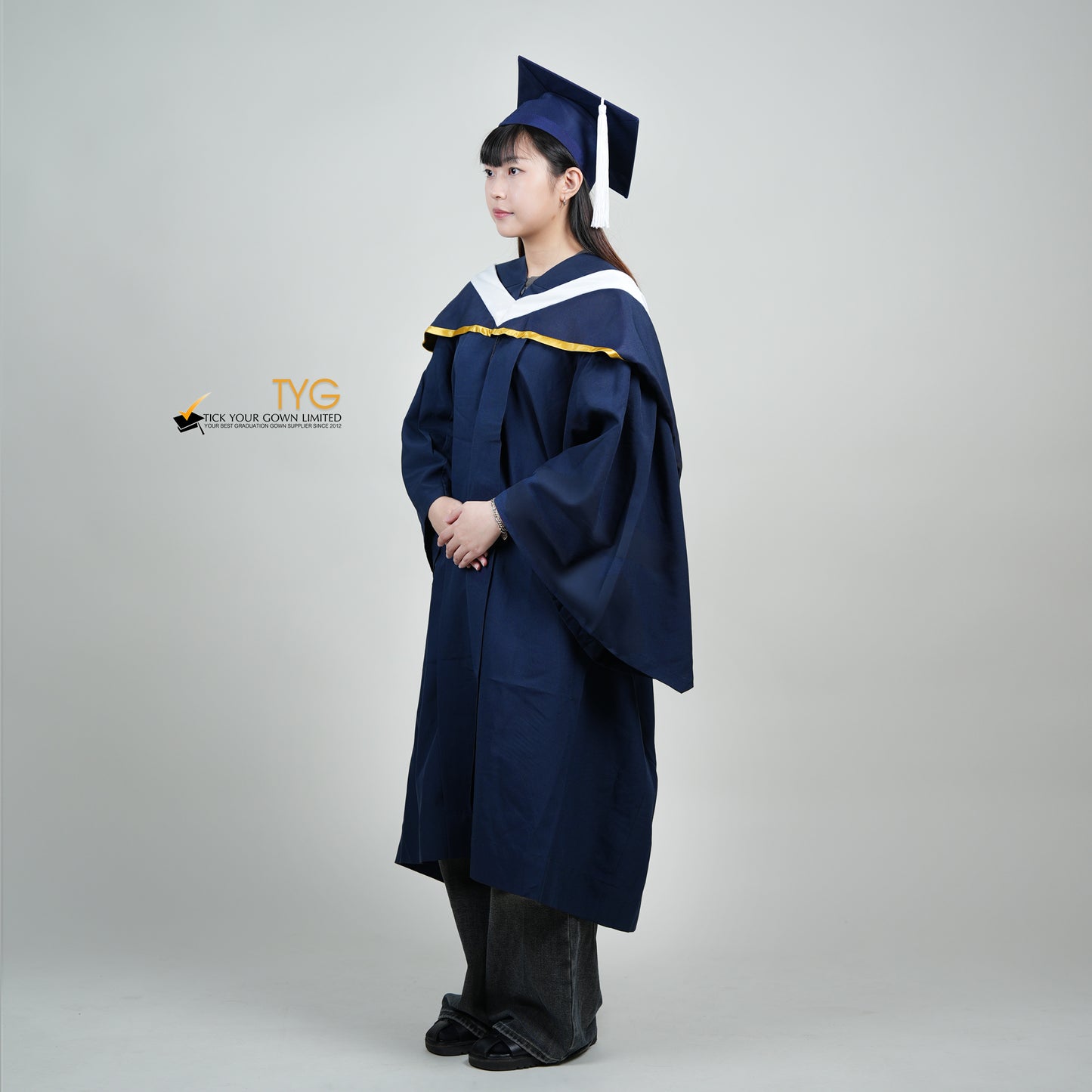 A full body profile view of a person in a navy blue graduation gown and a white hood with gold lining and with a mortarboard cap with a white tassel against a gray background.