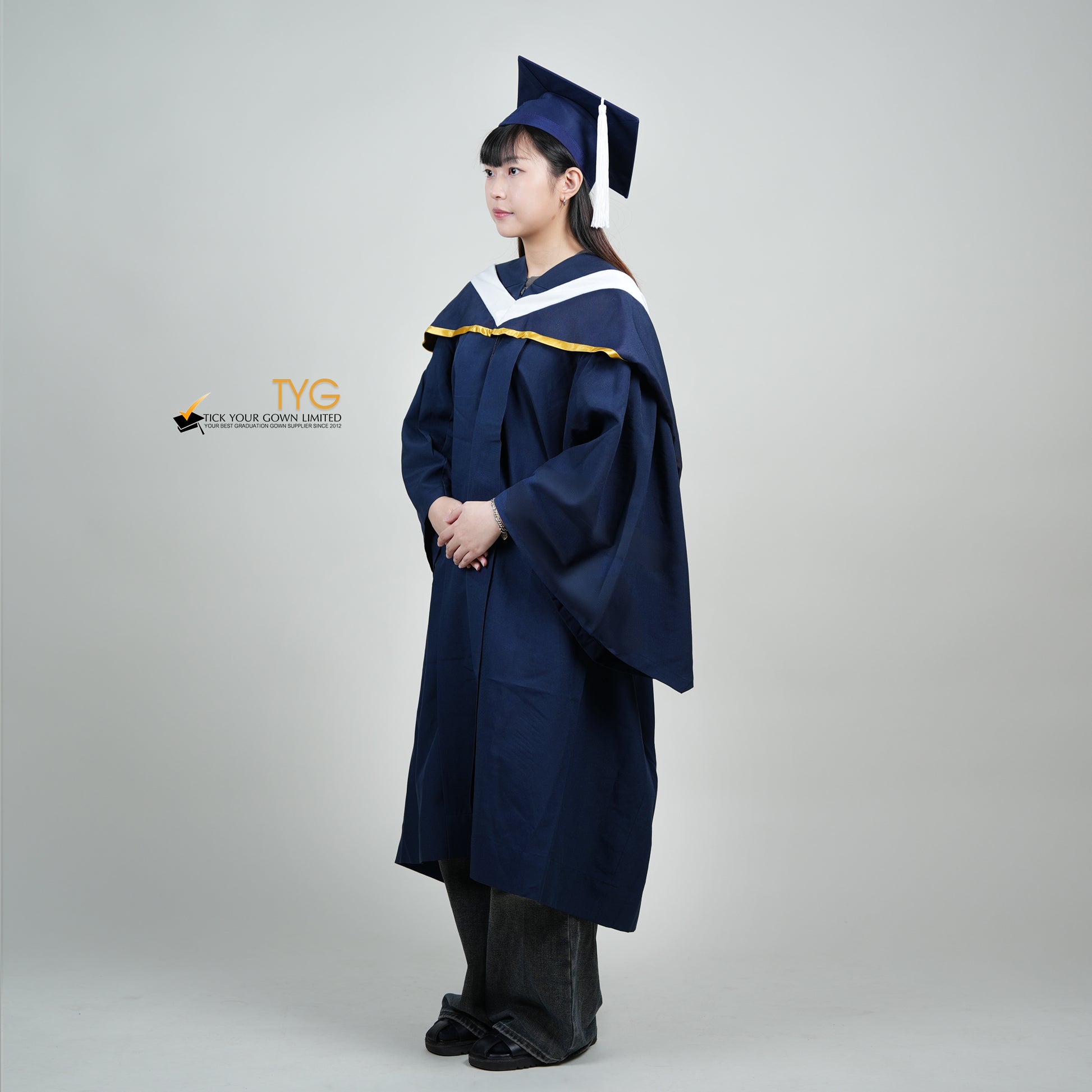 A full body profile view of a person in a navy blue graduation gown and a white hood with gold lining and with a mortarboard cap with a white tassel against a gray background.