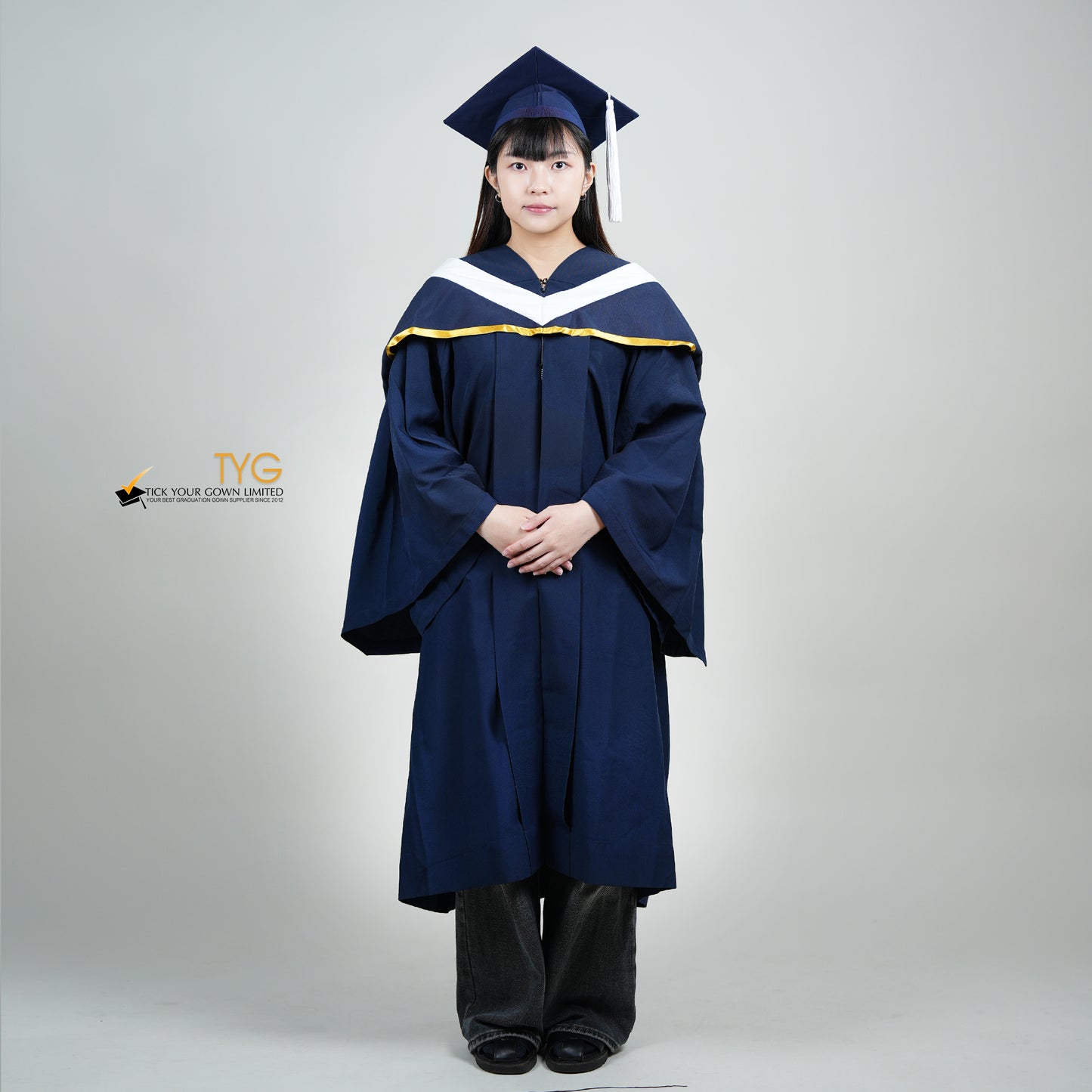 A full body shot of a person in a navy blue graduation gown and a white hood with gold lining and with a mortarboard cap with a white tassel against a gray background.