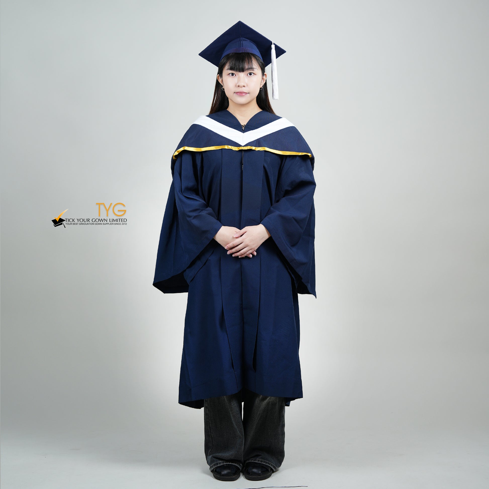 A full body shot of a person in a navy blue graduation gown and a white hood with gold lining and with a mortarboard cap with a white tassel against a gray background.
