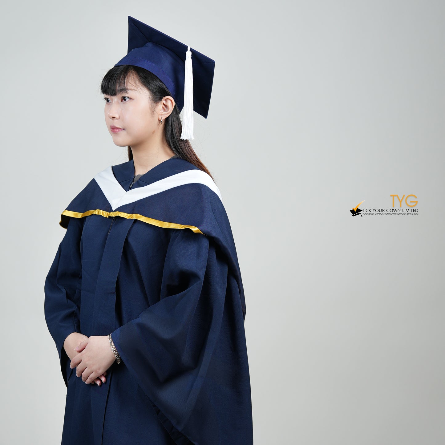 A profile view of a person in a navy blue graduation gown and a white hood with gold lining and with a mortarboard cap with a white tassel against a gray background.