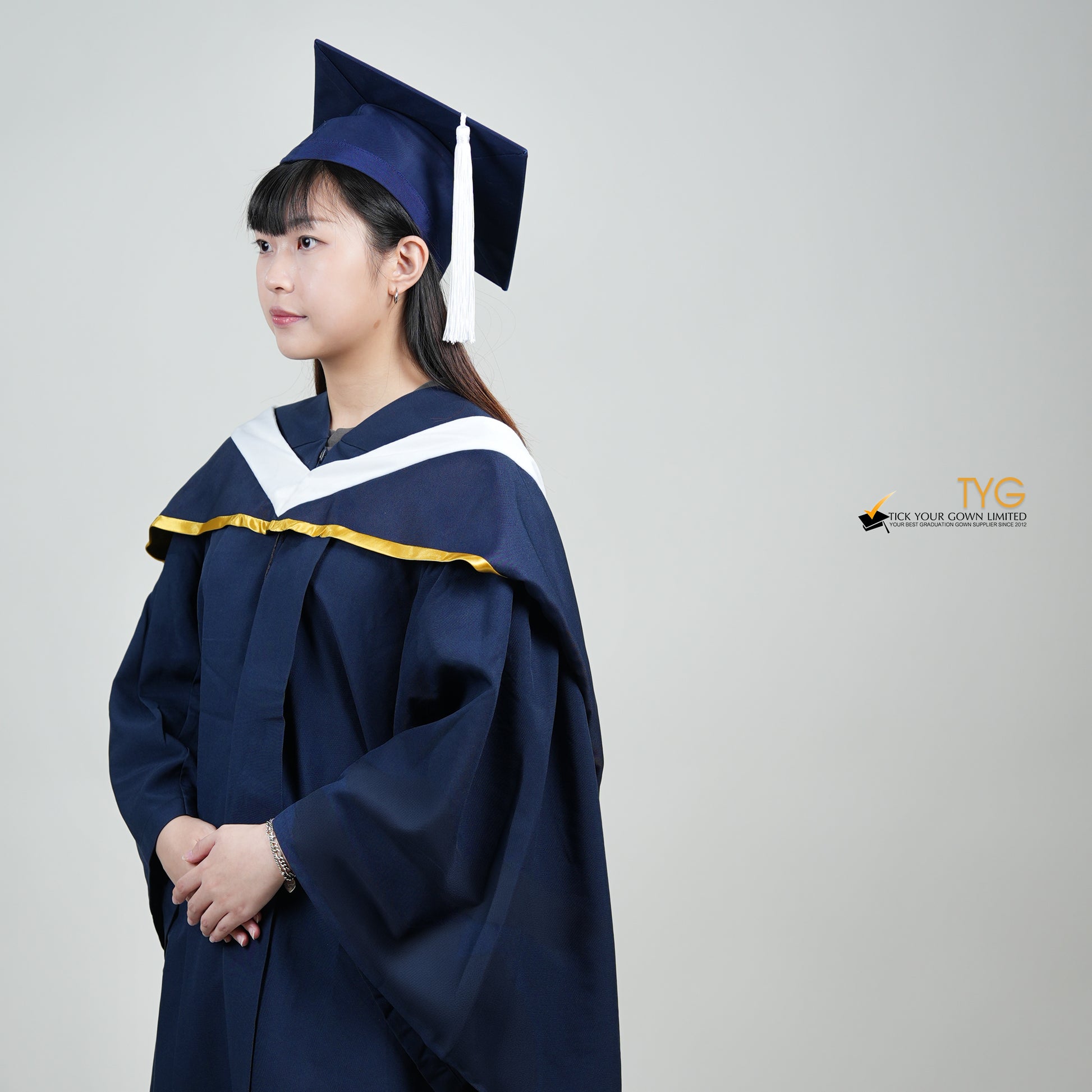 A profile view of a person in a navy blue graduation gown and a white hood with gold lining and with a mortarboard cap with a white tassel against a gray background.
