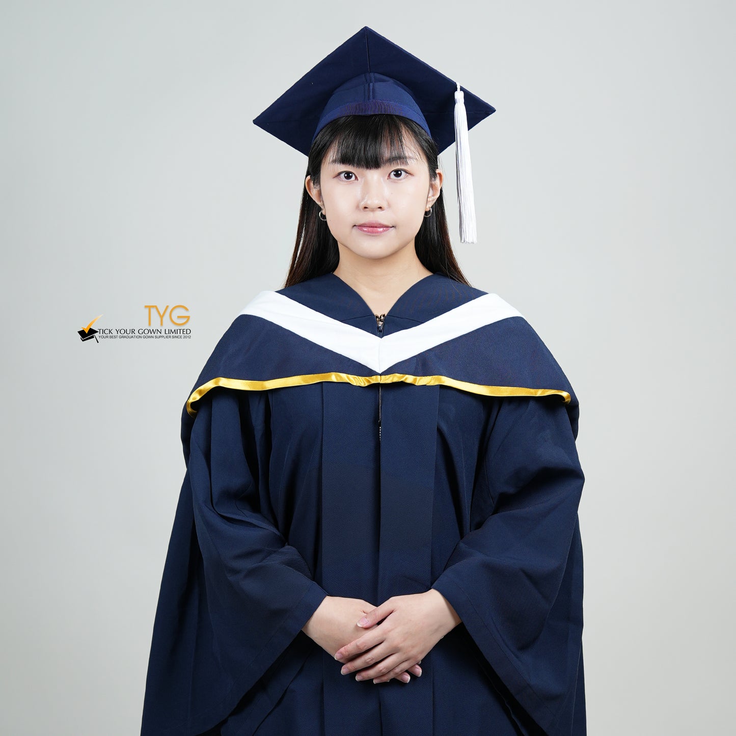 A person in a navy blue graduation gown and a white hood with gold lining and with a mortarboard cap with a white tassel against a gray background.