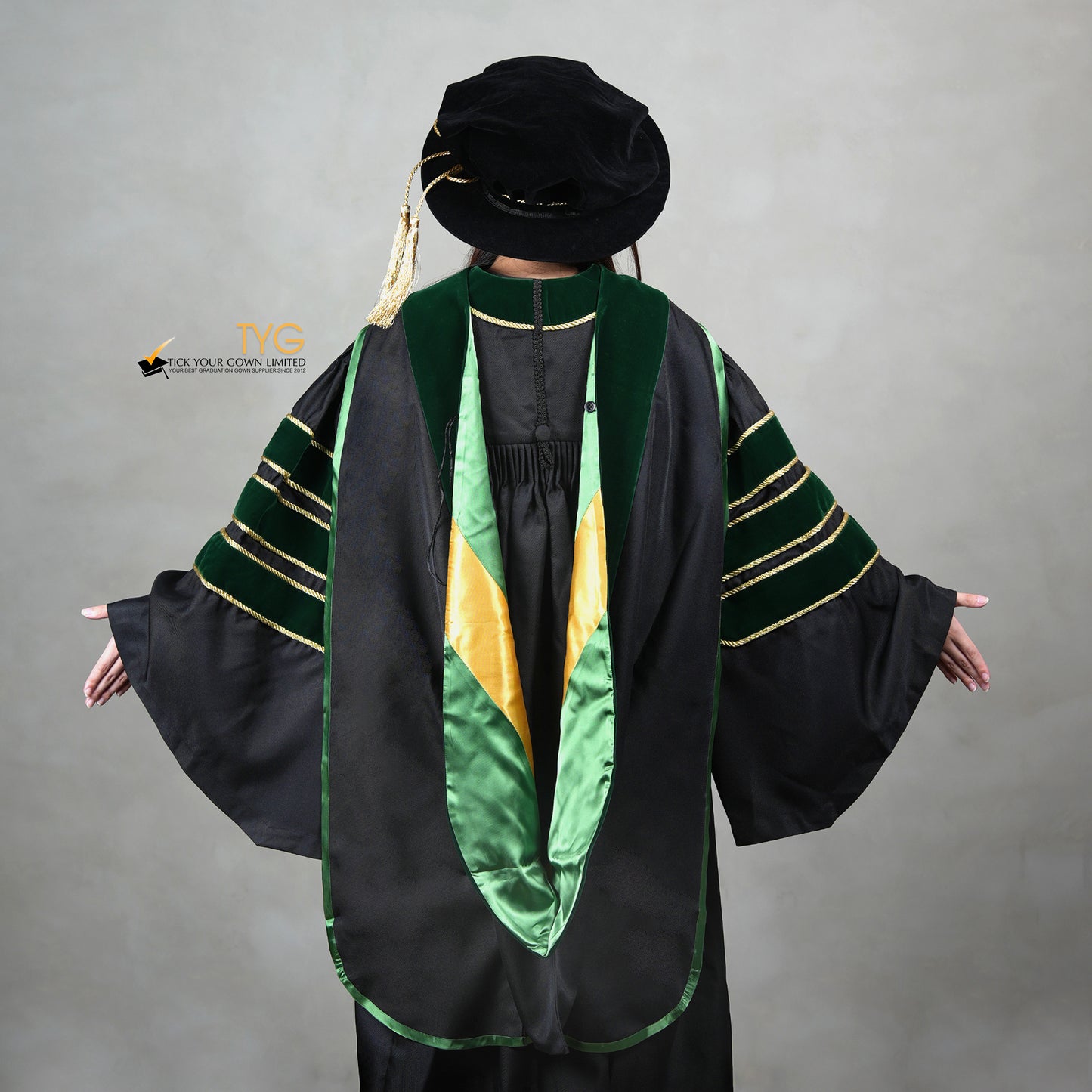 A back view of a person wearing a PhD graduation gown in black with forest green velvet panels, matching sleeve bars, gold trim, and a matching doctoral bonnet with gold tassel.