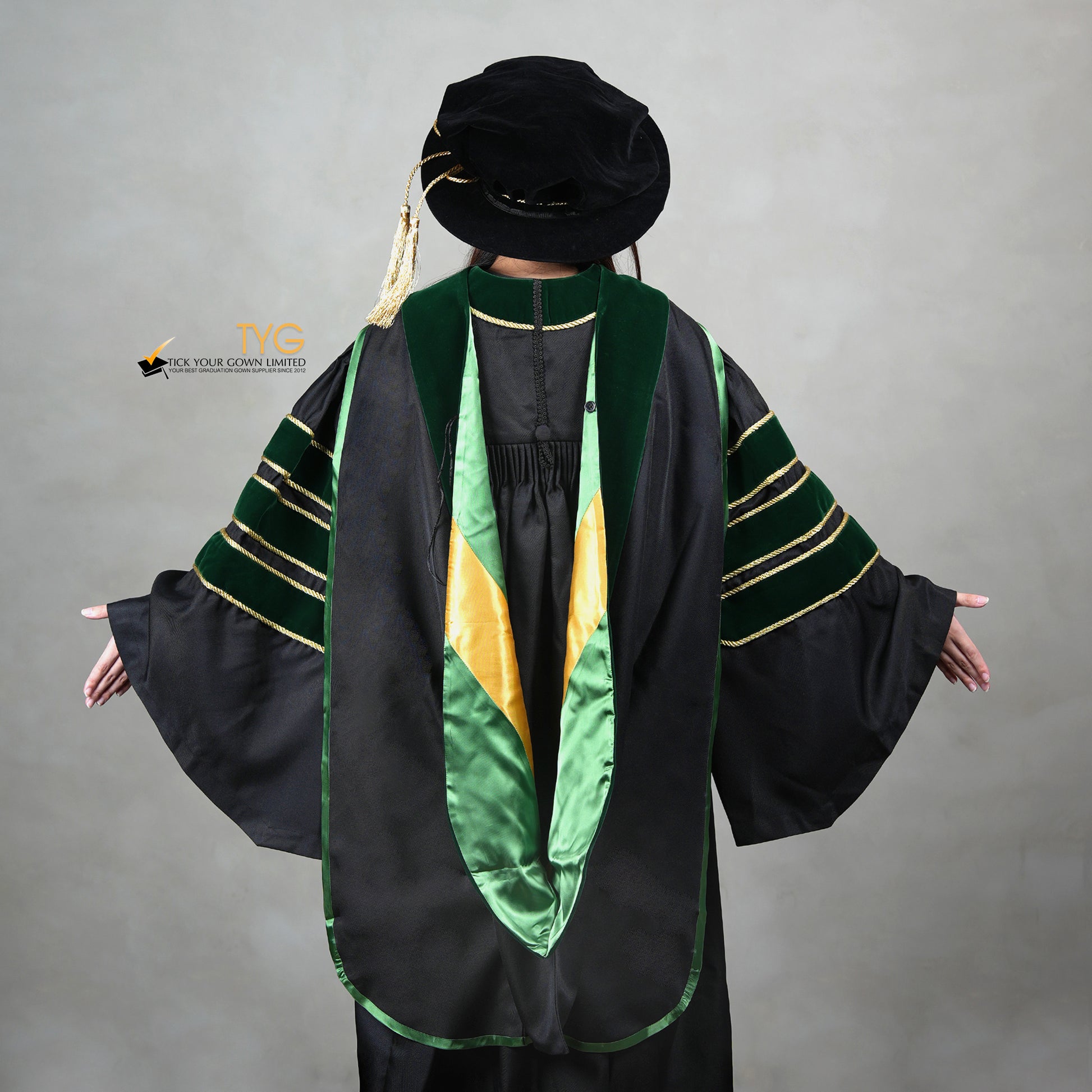 A back view of a person wearing a PhD graduation gown in black with forest green velvet panels, matching sleeve bars, gold trim, and a matching doctoral bonnet with gold tassel.