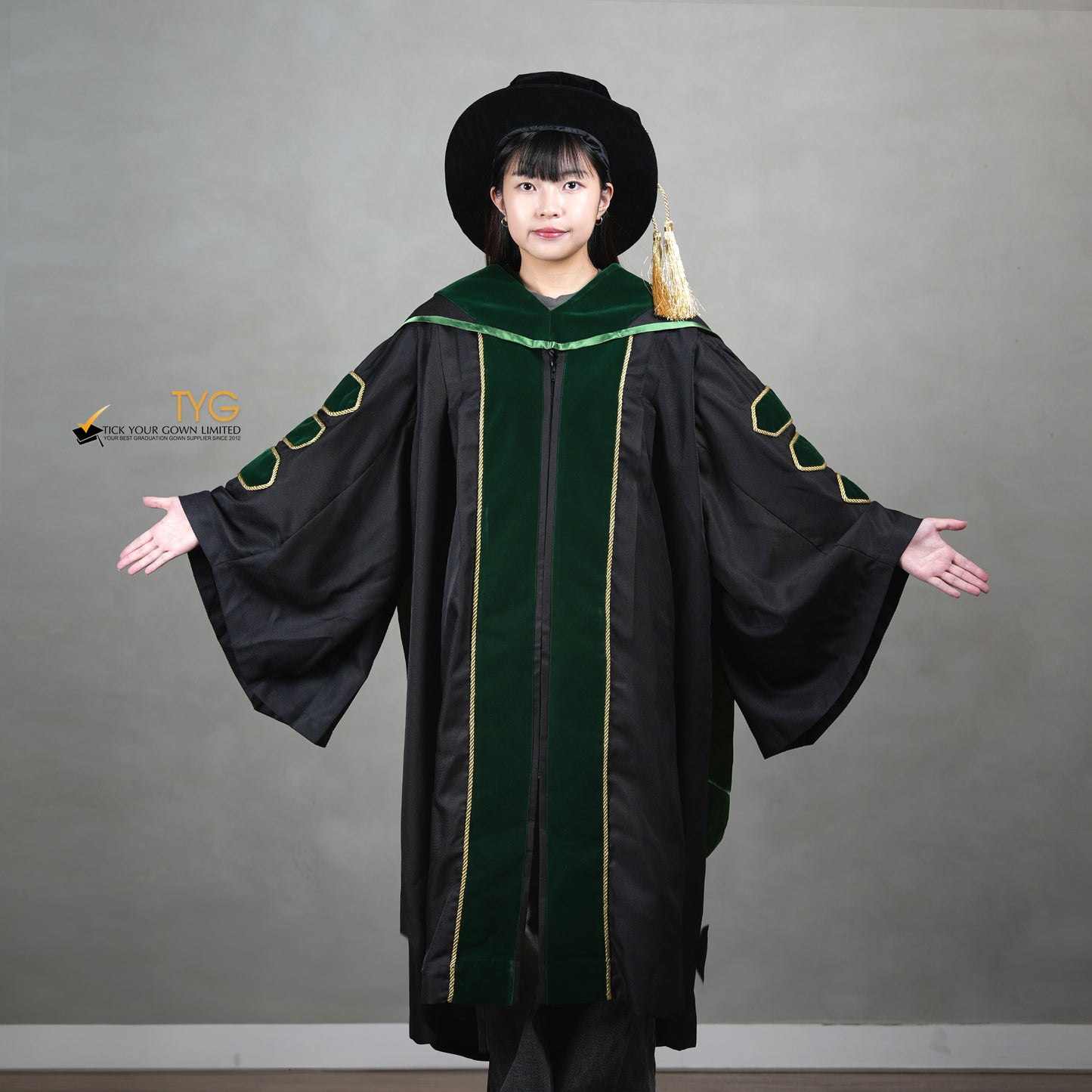 A showcase front view of a person wearing a PhD graduation gown in black with forest green velvet panels, matching sleeve bars, gold trim, and a matching doctoral bonnet with gold tassel.