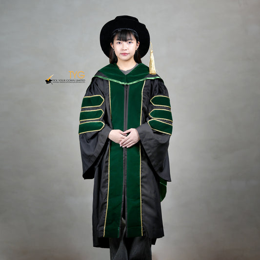 A front view of a person wearing a PhD graduation gown in black with forest green velvet panels, matching sleeve bars, gold trim, and a matching doctoral bonnet with gold tassel.