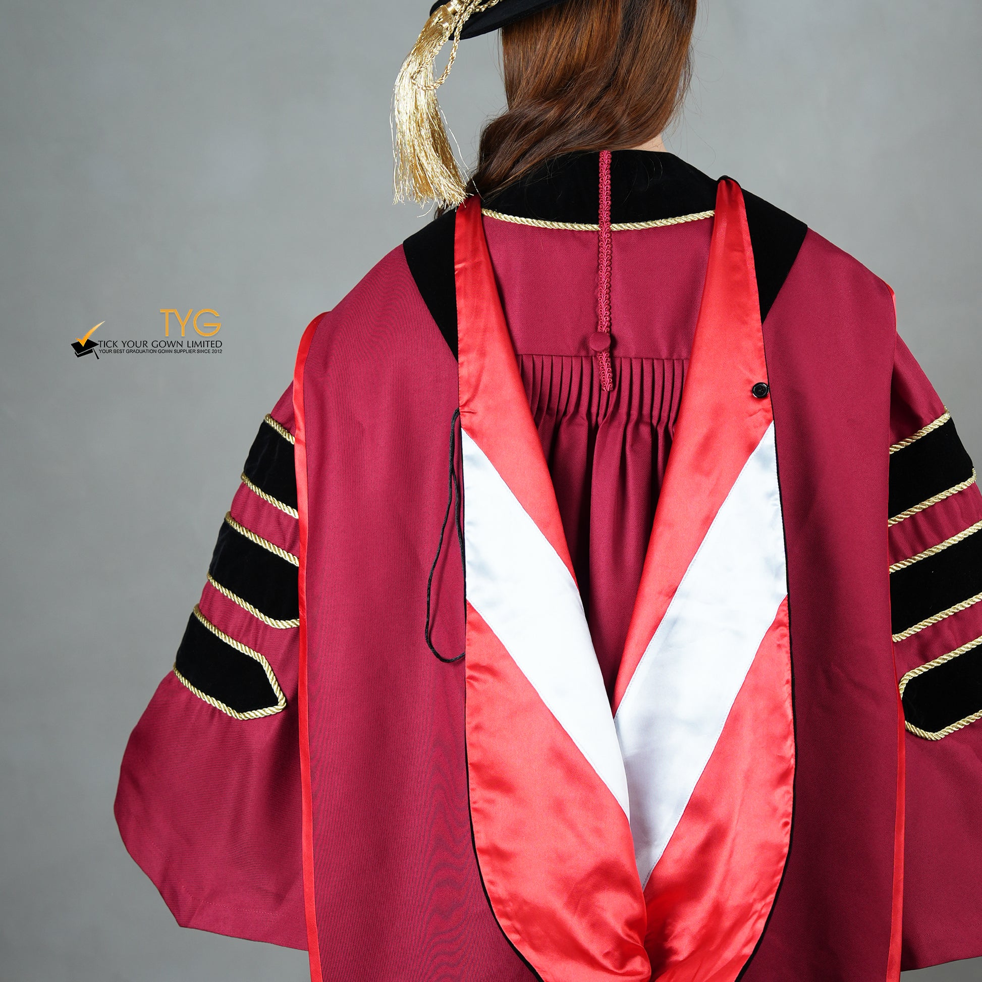 A detailed back view  of a person wearing a doctoral graduation gown in maroon with black velvet panels, matching black velvet sleeve bars, gold trim, and a black doctoral bonnet with gold tassel.