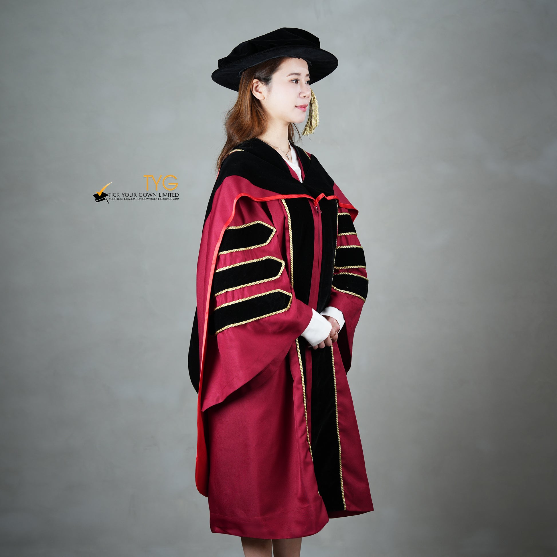 A profile view of a person wearing a doctoral graduation gown in maroon with black velvet panels, matching black velvet sleeve bars, gold trim, and a black doctoral bonnet with gold tassel.