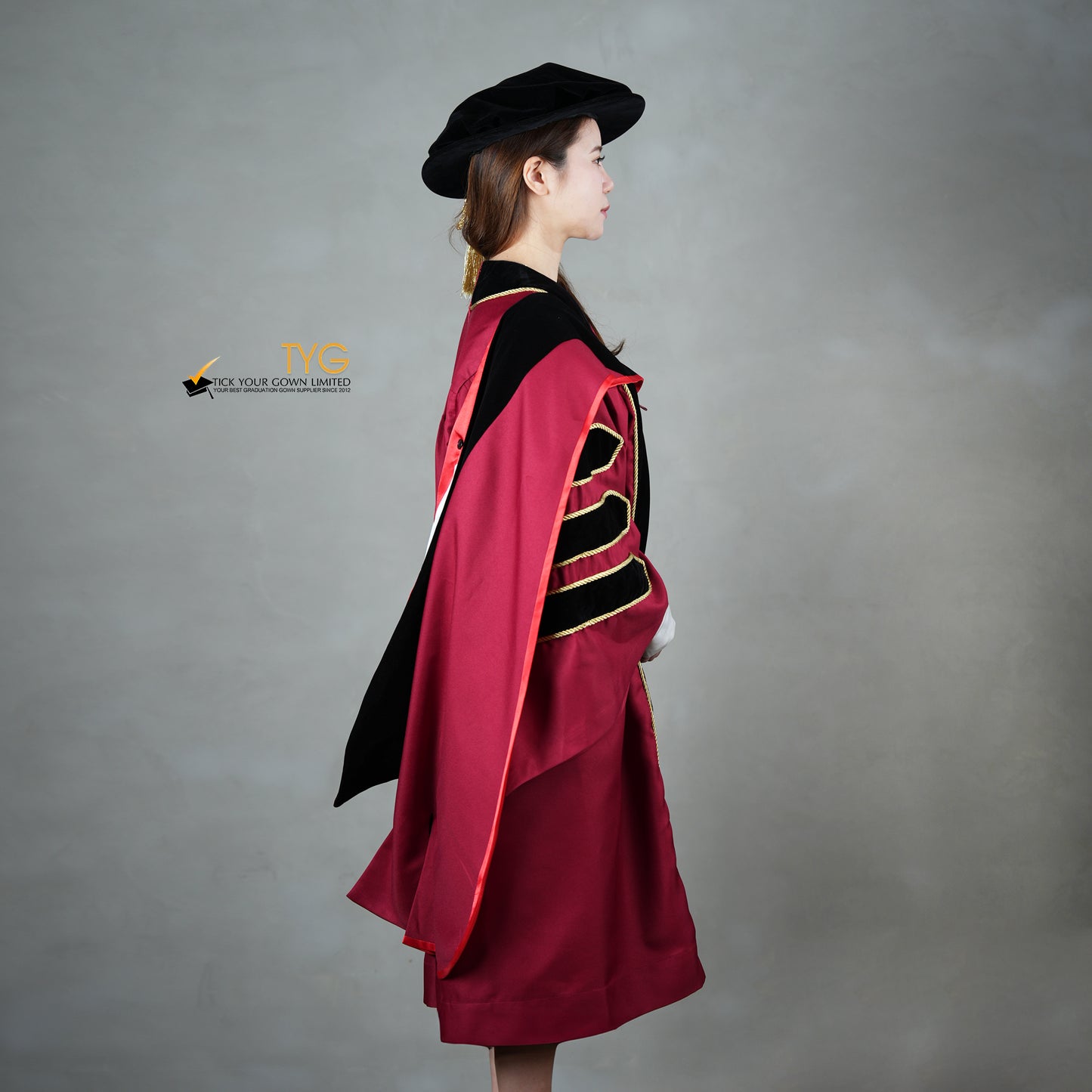 A side view  of a person wearing a doctoral graduation gown in maroon with black velvet panels, matching black velvet sleeve bars, gold trim, and a black doctoral bonnet with gold tassel.