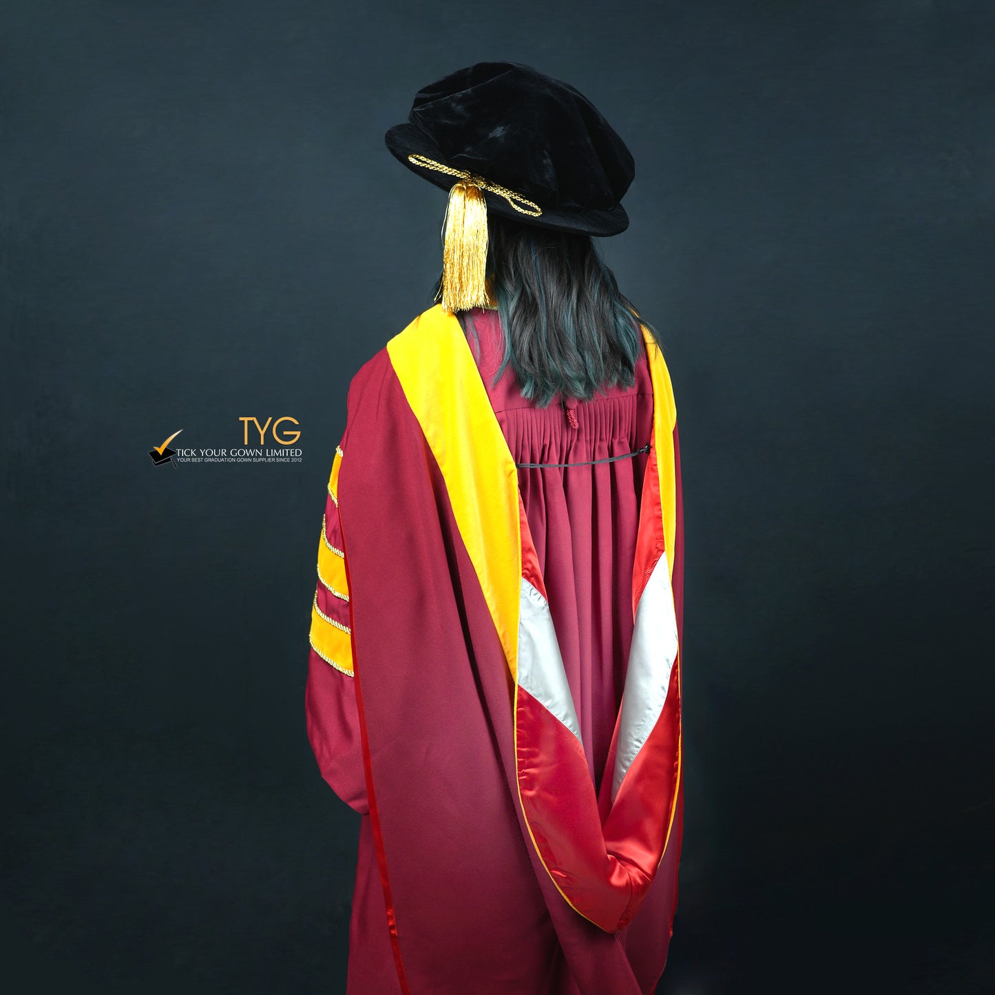 A back profile view of a person wearing a classic PhD graduation gown in maroon with yellow velvet panels, matching sleeve bars, and a black doctoral bonnet with gold tassel