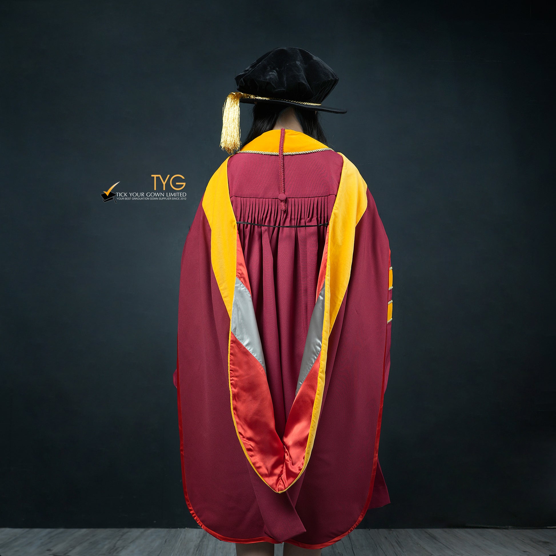 Back view of a person wearing a classic PhD graduation gown in maroon with yellow velvet panels, matching sleeve bars, and a black doctoral bonnet with gold tassel