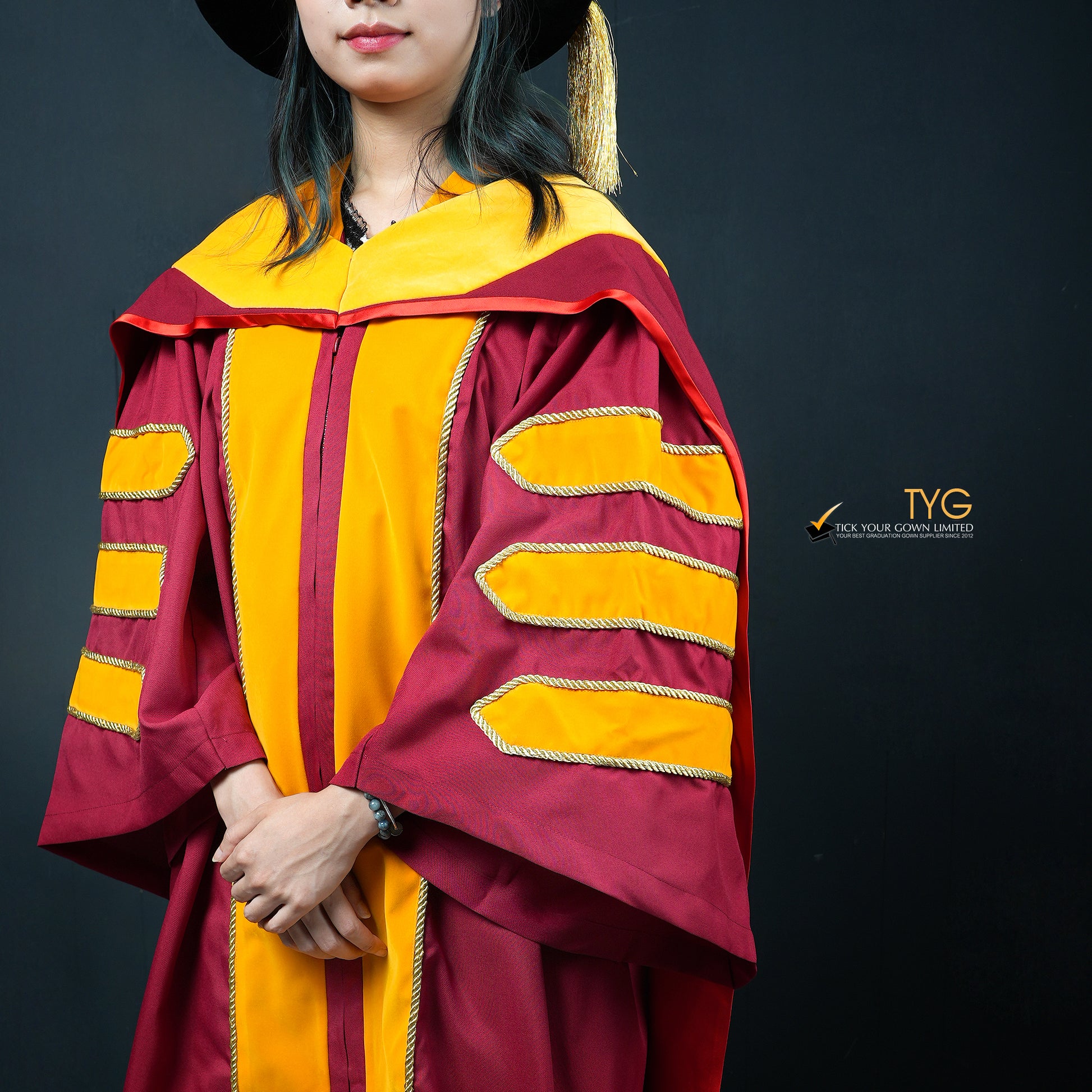A detailed profile view of a person wearing a classic PhD graduation gown in maroon with yellow velvet panels, matching sleeve bars, and a black doctoral bonnet with gold tassel