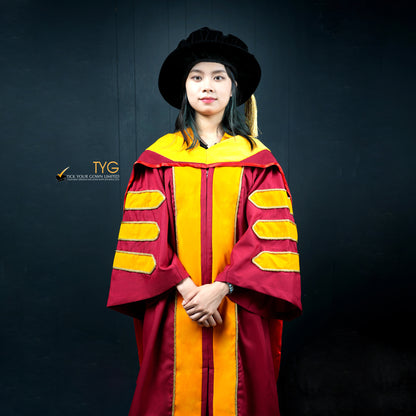 Front view of a person wearing a classic PhD graduation gown in maroon with yellow velvet panels, matching sleeve bars, and a black doctoral bonnet with gold tassel
