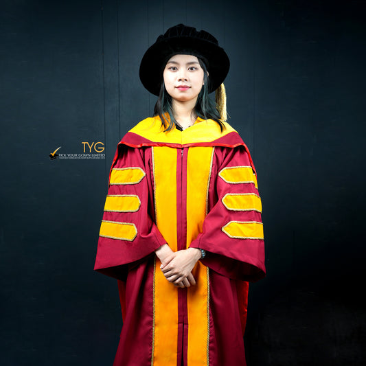 Front view of a person wearing a classic PhD graduation gown in maroon with yellow velvet panels, matching sleeve bars, and a black doctoral bonnet with gold tassel
