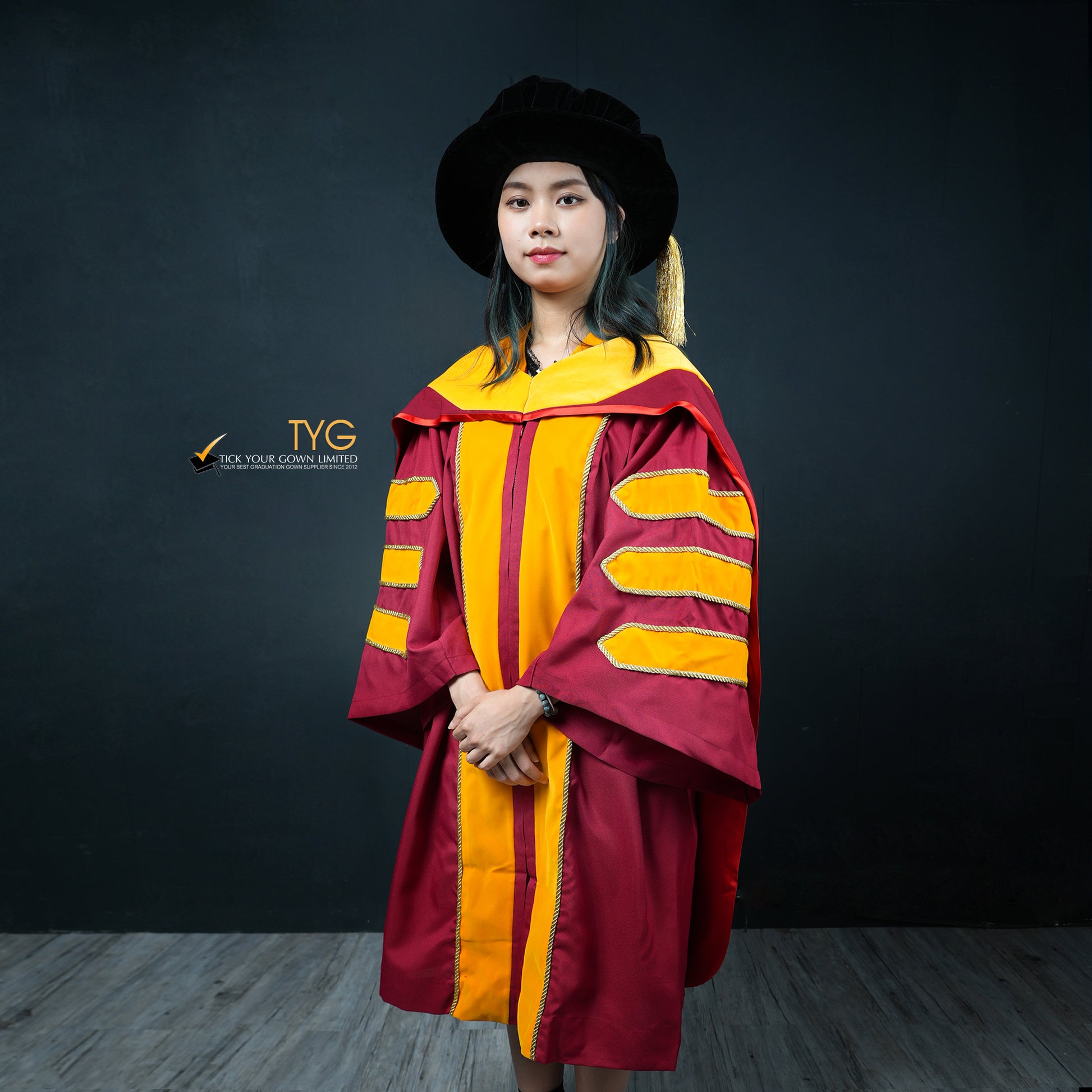 A profile view of a person wearing a classic PhD graduation gown in maroon with yellow velvet panels, matching sleeve bars, and a black doctoral bonnet with gold tassel