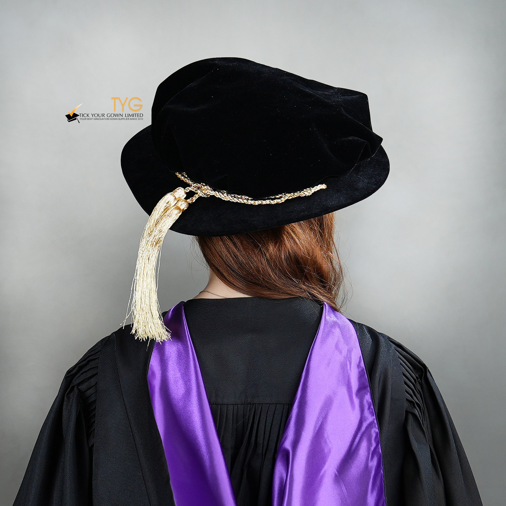 Back view of a person wearing a black doctoral bonnet and purple gown with a gold tassel on a gray background