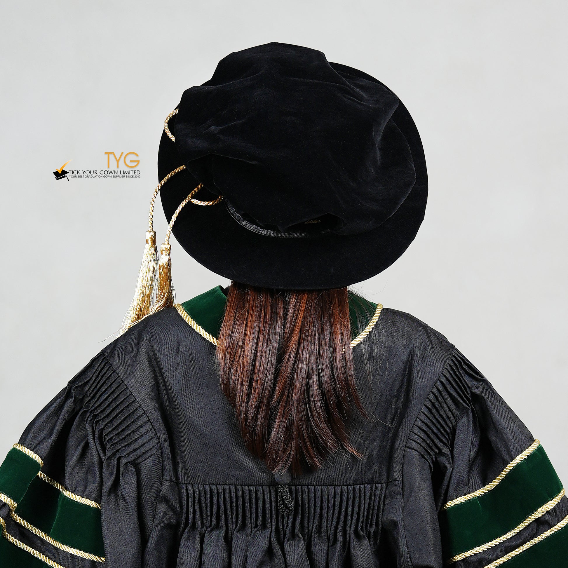 Rear view of a person wearing a black doctoral bonnet and gown with gold accents on a light gray background.