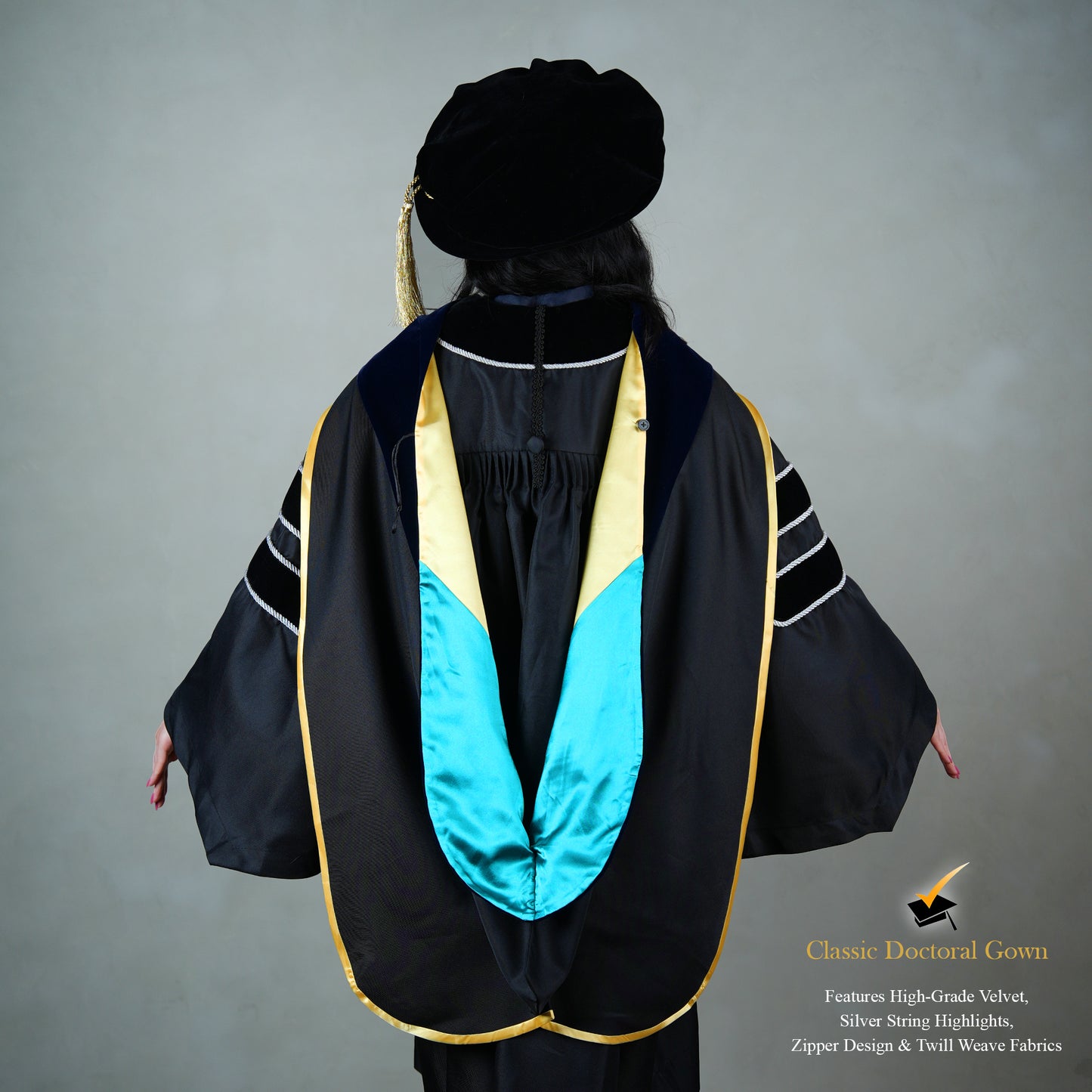 Back view of a person wearing a PhD classic black velvet graduation gown with silver sleeve bars, silver front trim, and a black doctoral bonnet with gold tassel.