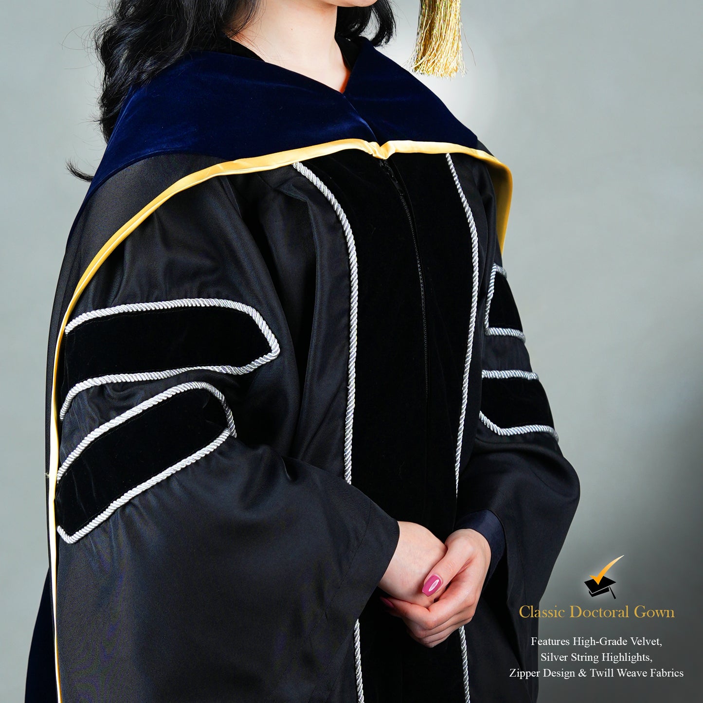 Detailed profile view of a person wearing a PhD classic black velvet graduation gown with silver sleeve bars, silver front trim, and a black doctoral bonnet with gold tassel.