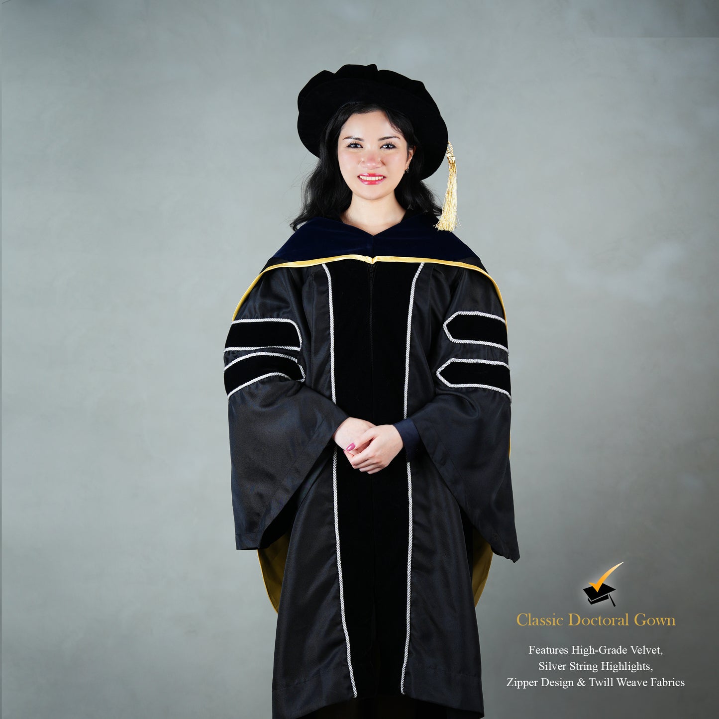 Front view of a person wearing a PhD classic black velvet graduation gown with silver sleeve bars, silver front trim, and a black doctoral bonnet with gold tassel.