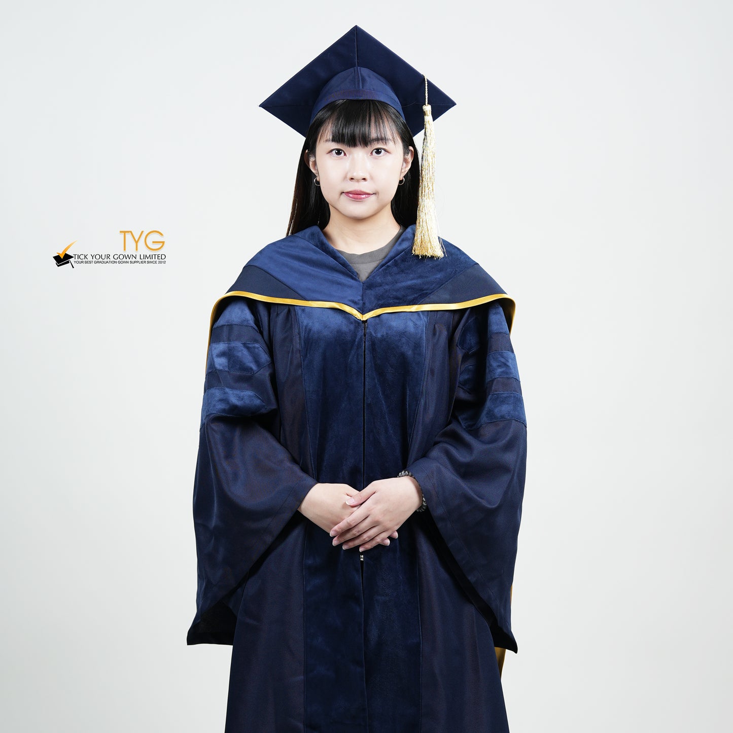 A detailed front view of a person wearing a PhD classic navy blue velvet graduation gown with yellow lining hood, and a navy blue doctoral bonnet with gold tassel.