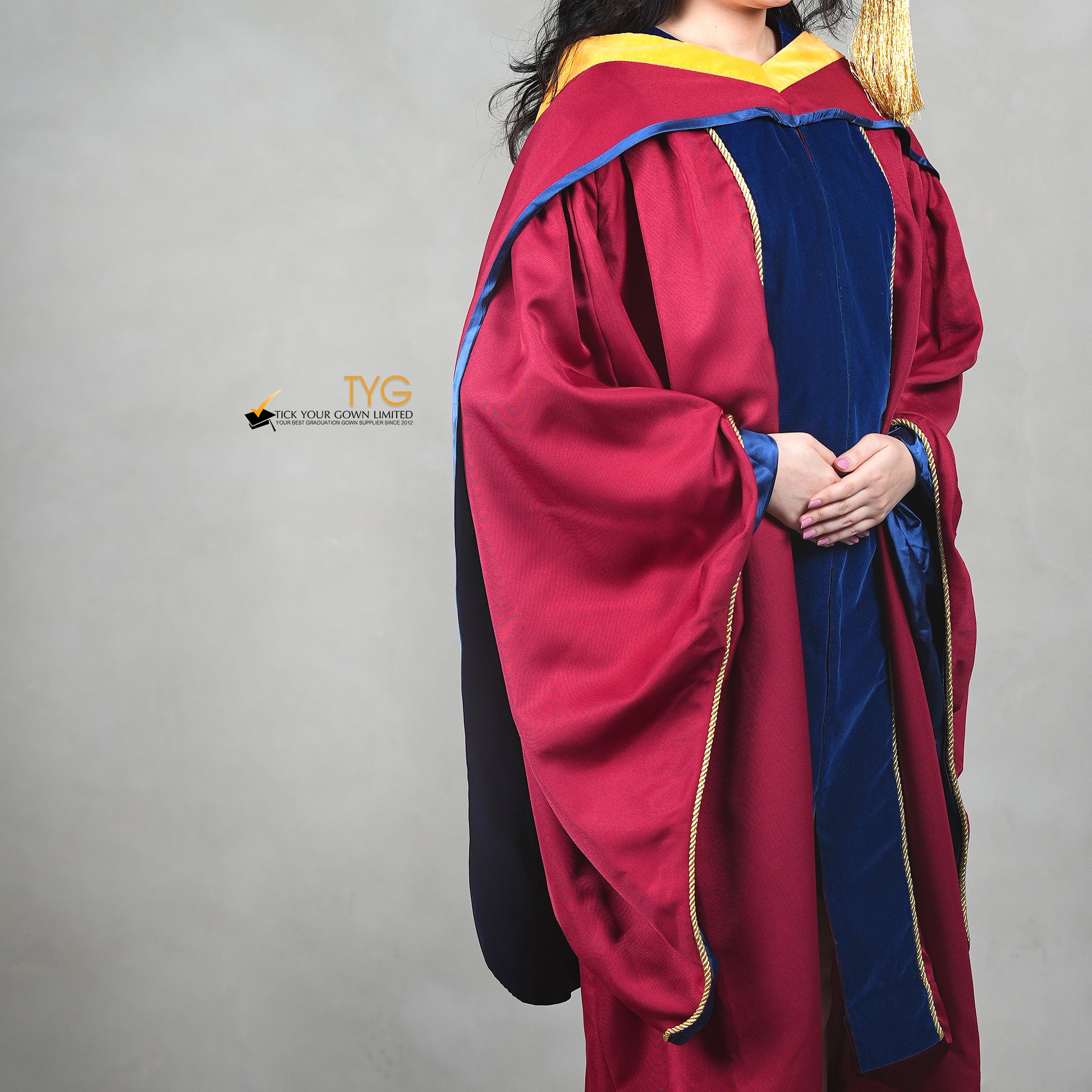 A profile view of a person wearing a premium honorary graduation gown in maroon with royal blue velvet panels, gold trim, and a black doctoral bonnet with gold tassel