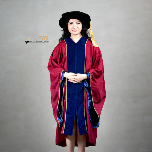 Front view of a person wearing a premium honorary graduation gown in maroon with royal blue velvet panels, gold trim, and a matching doctoral bonnet with gold tassel.