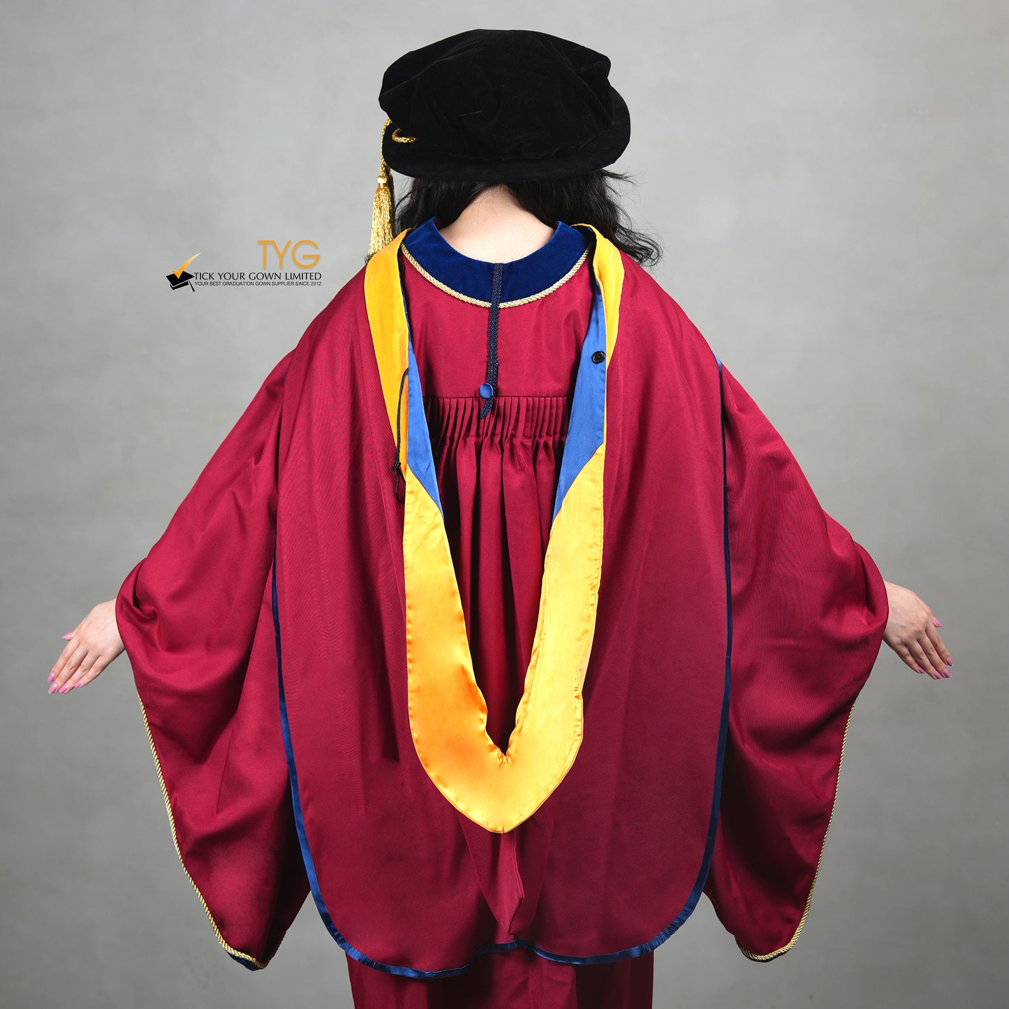 A back view of a person showcasing a premium honorary graduation gown in maroon with royal blue velvet panels, gold trim, and a black doctoral bonnet with gold tassel