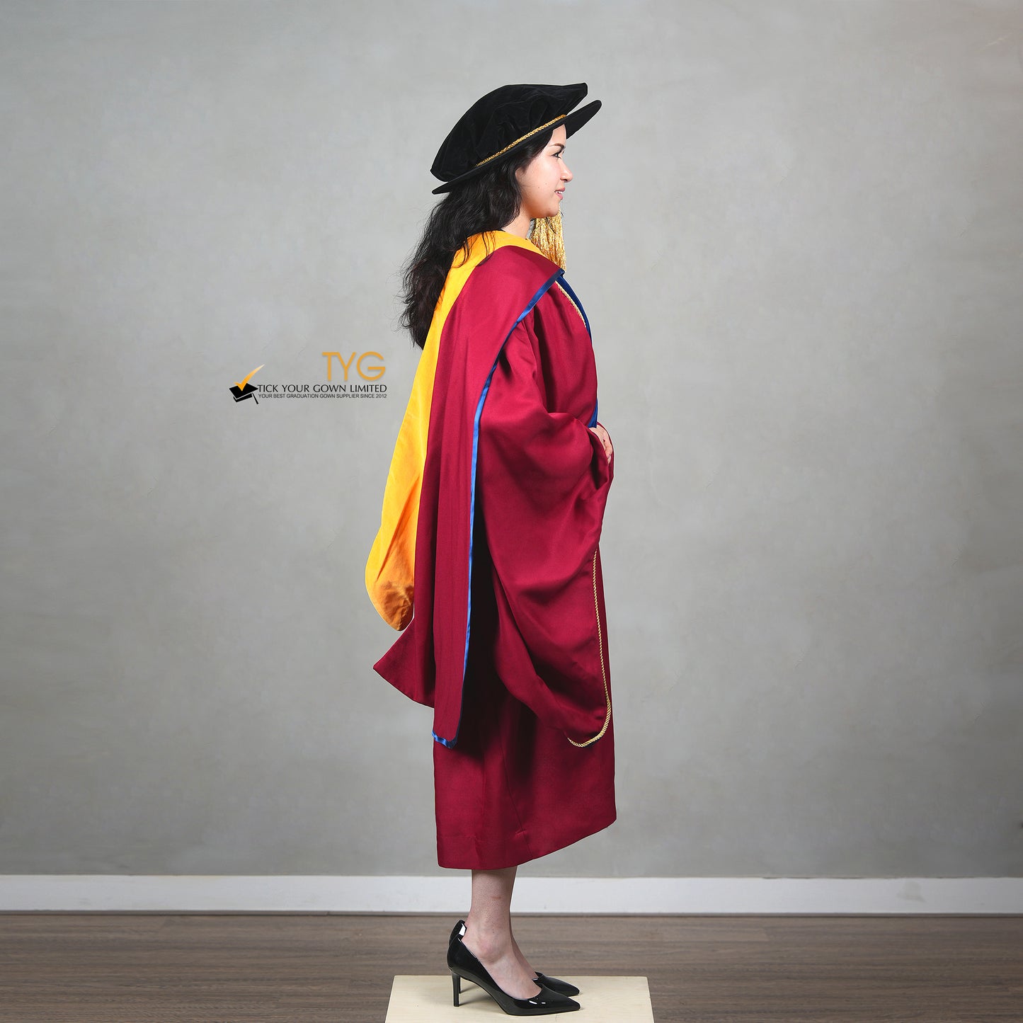 A side view of a person wearing a premium honorary graduation gown in maroon with royal blue velvet panels, gold trim, and a black doctoral bonnet with gold tassel