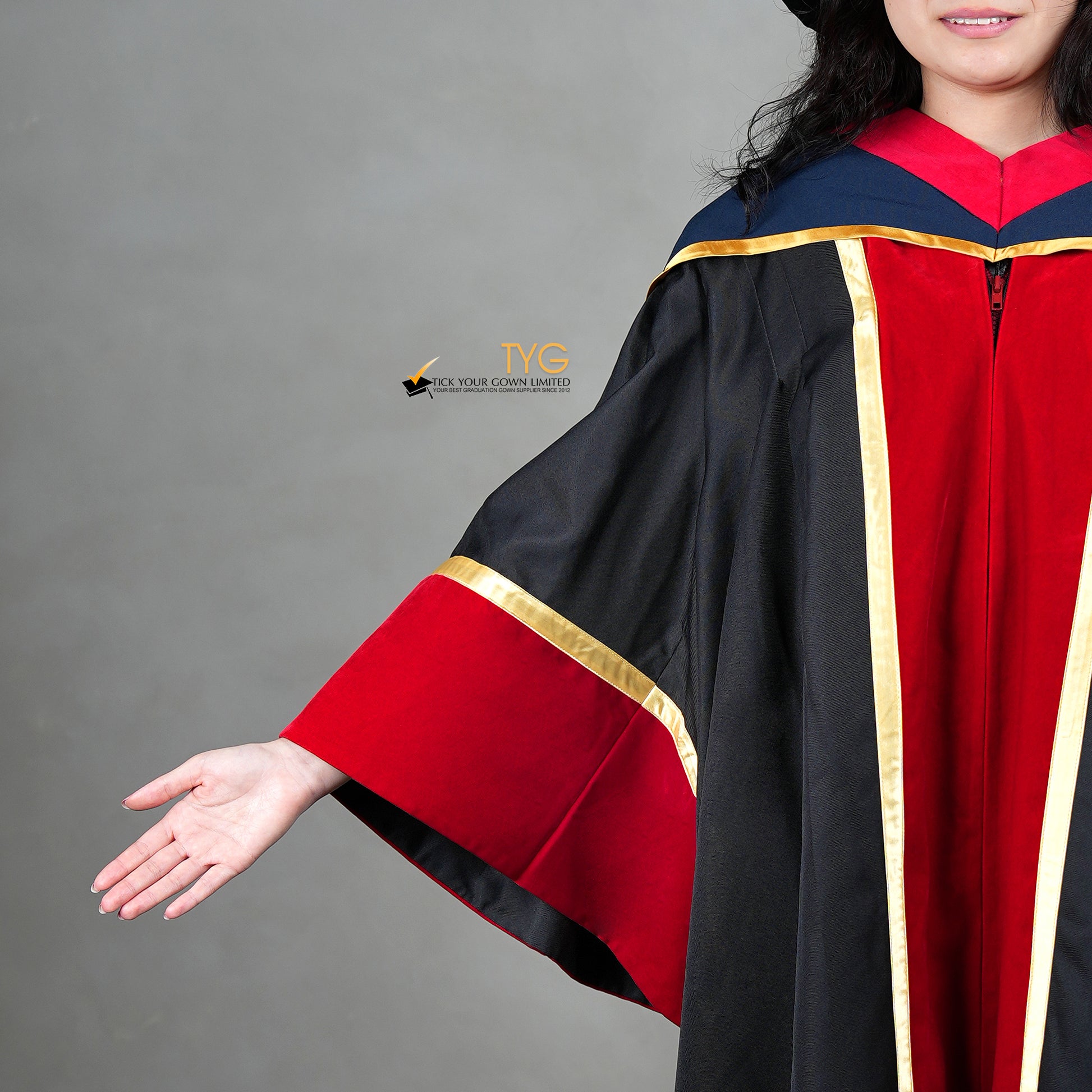 A detailed front view of a person wearing a premium honorary graduation gown in black with red velvet panels, gold trim, matching red velvet sleeve cuffs, and a doctoral bonnet with gold tassel