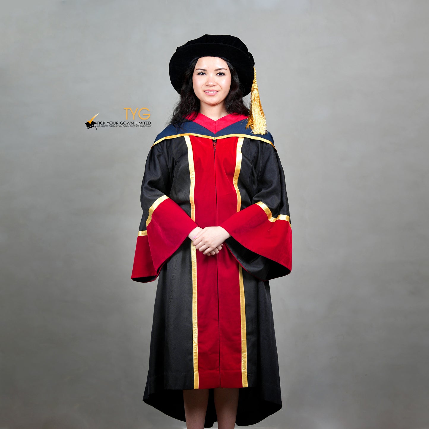Front view of a person wearing a premium honorary graduation gown in black with red velvet panels, gold trim, matching red velvet sleeve cuffs, and a doctoral bonnet with gold tassel