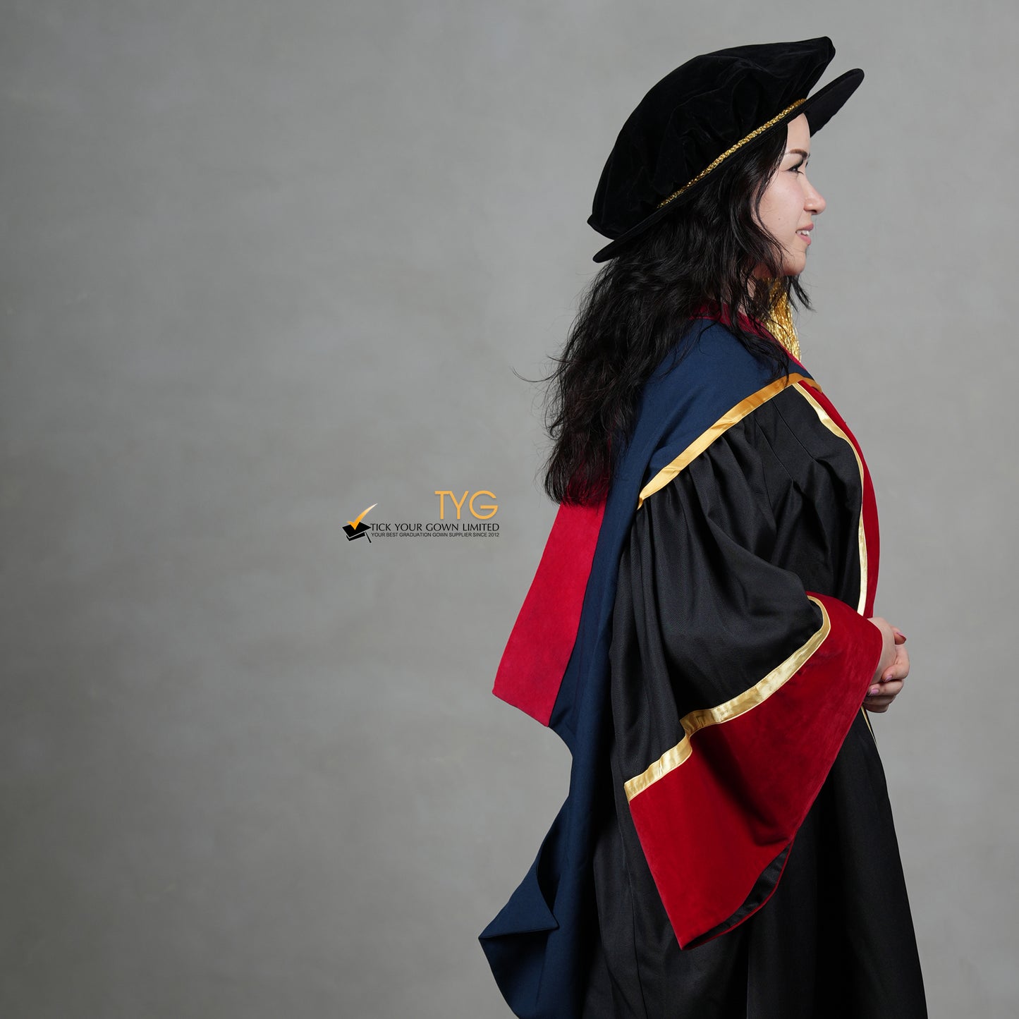 A side view of a person wearing a premium honorary graduation gown in black with red velvet panels, gold trim, matching red velvet sleeve cuffs, and a doctoral bonnet with gold tassel