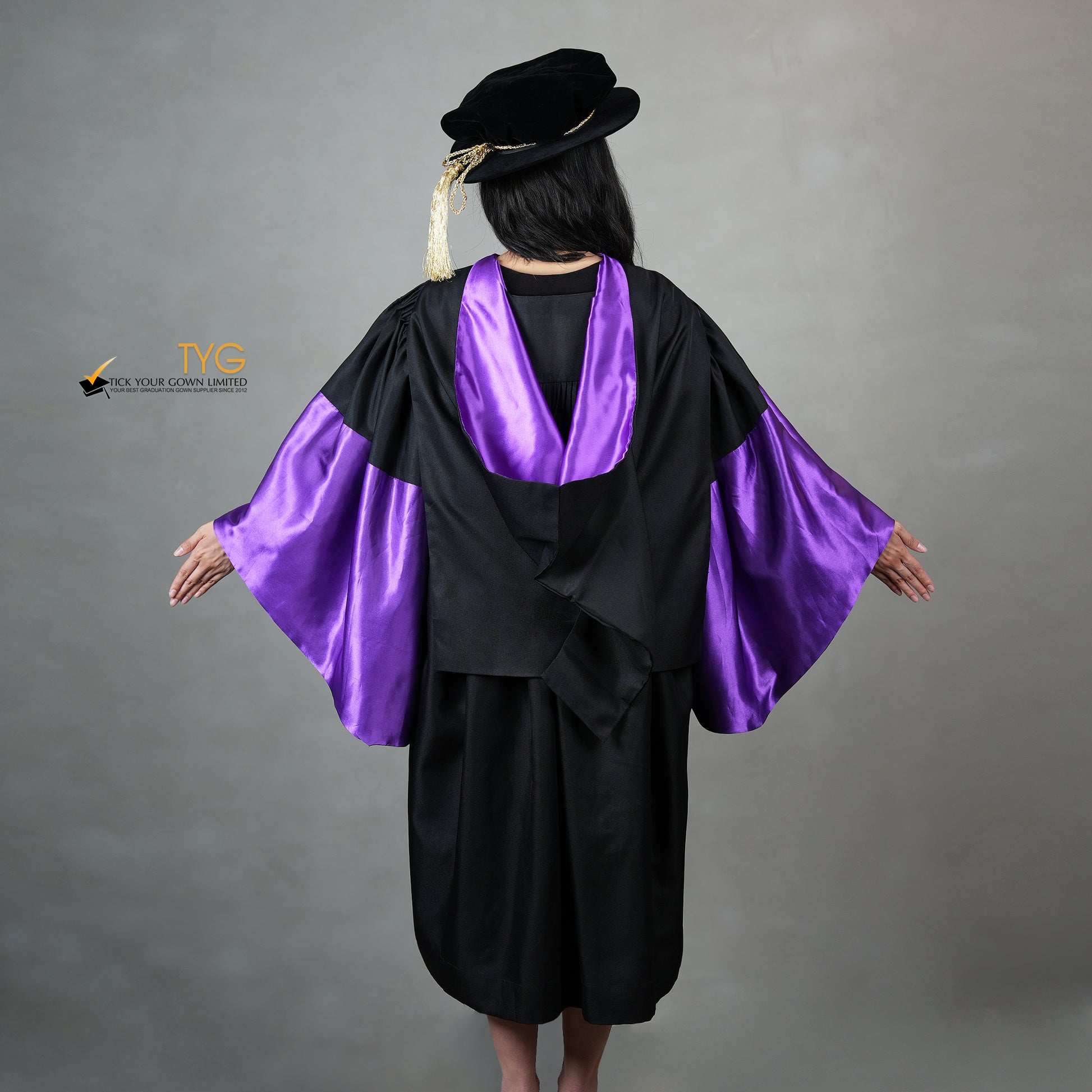 A back view of a person wearing a graduation gown in black with purple satin panels, matching purple satin sleeve cuffs, and a black doctoral bonnet with gold tassel.