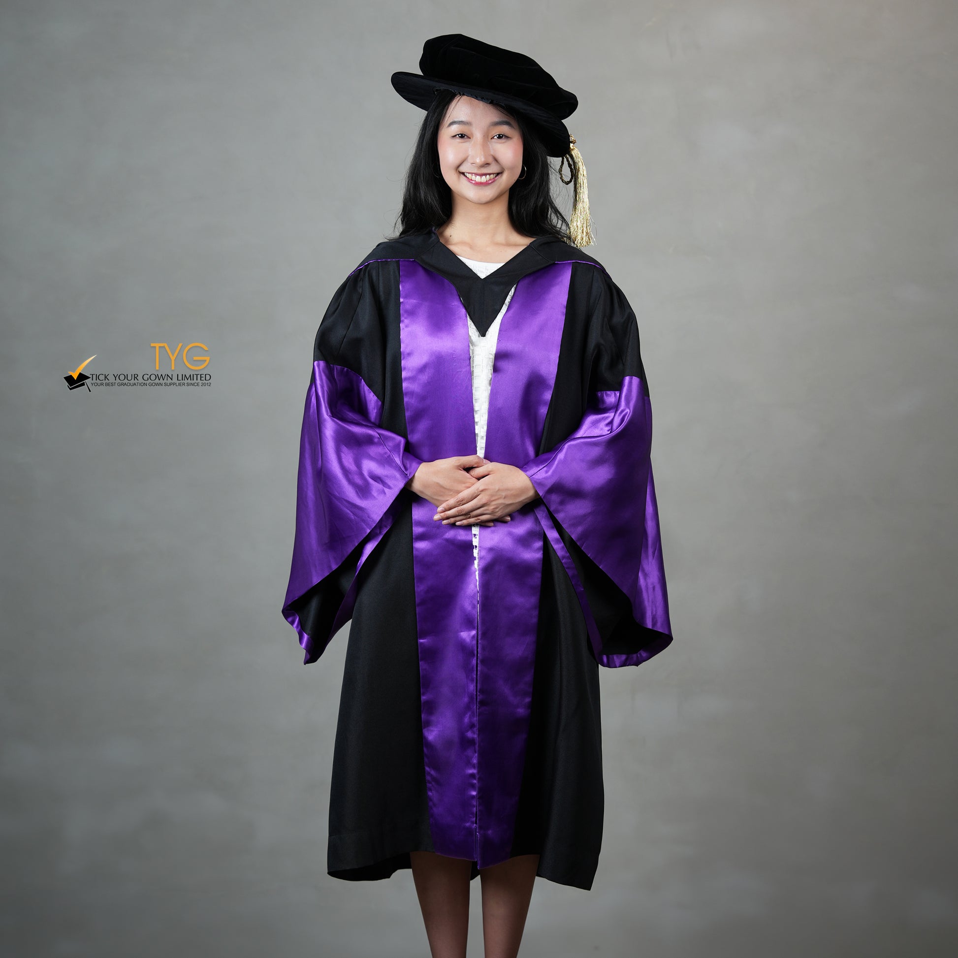 A front view of a person wearing a graduation gown in black with purple satin panels, matching purple satin sleeve cuffs, and a black doctoral bonnet with gold tassel.