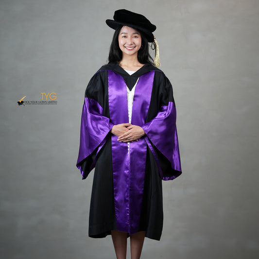 A front view of a person wearing a graduation gown in black with purple satin panels, matching purple satin sleeve cuffs, and a black doctoral bonnet with gold tassel.