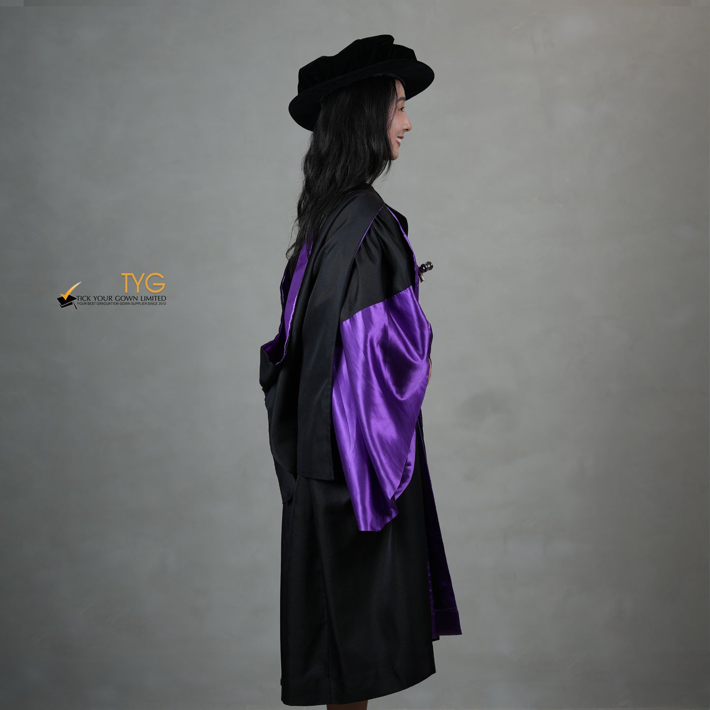 A side view of a person wearing a graduation gown in black with purple satin panels, matching purple satin sleeve cuffs, and a black doctoral bonnet with gold tassel.