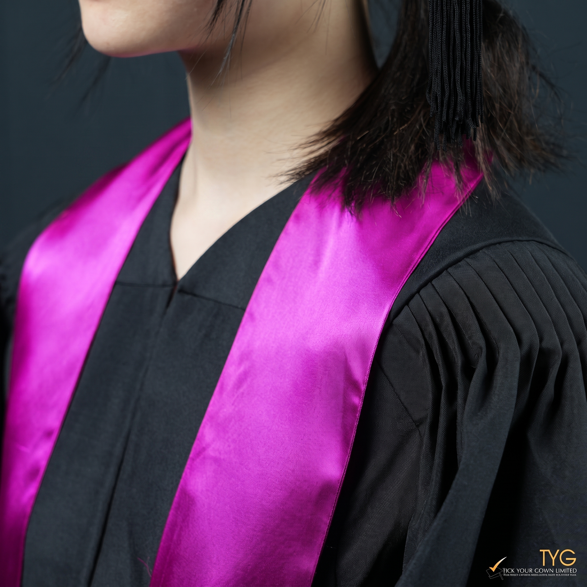 A closeup view of a person wearing a black graduation gown with a pink graduation sash stole on a dark background