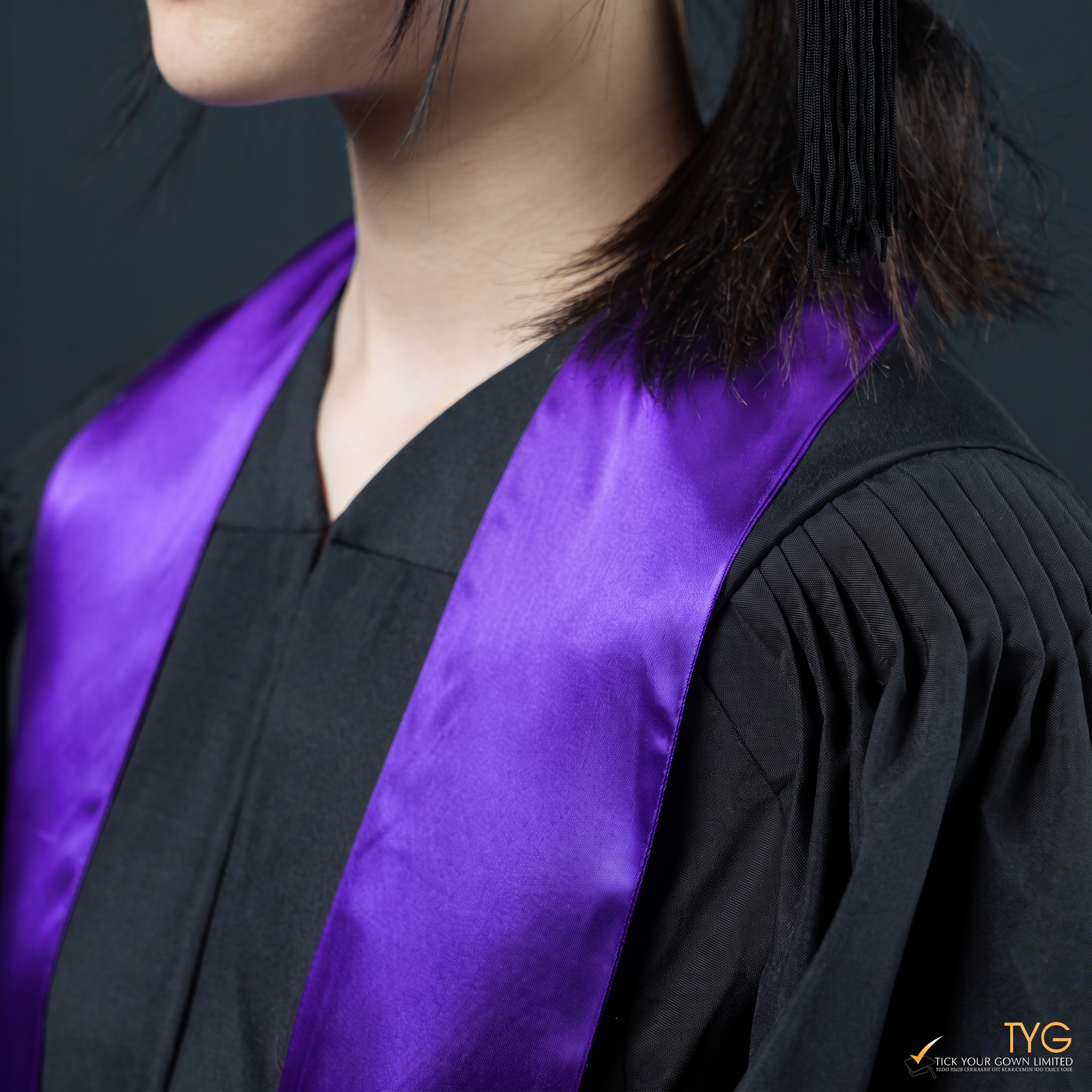 Person wearing a black graduation gown with a purple graduation sash stole on a dark background